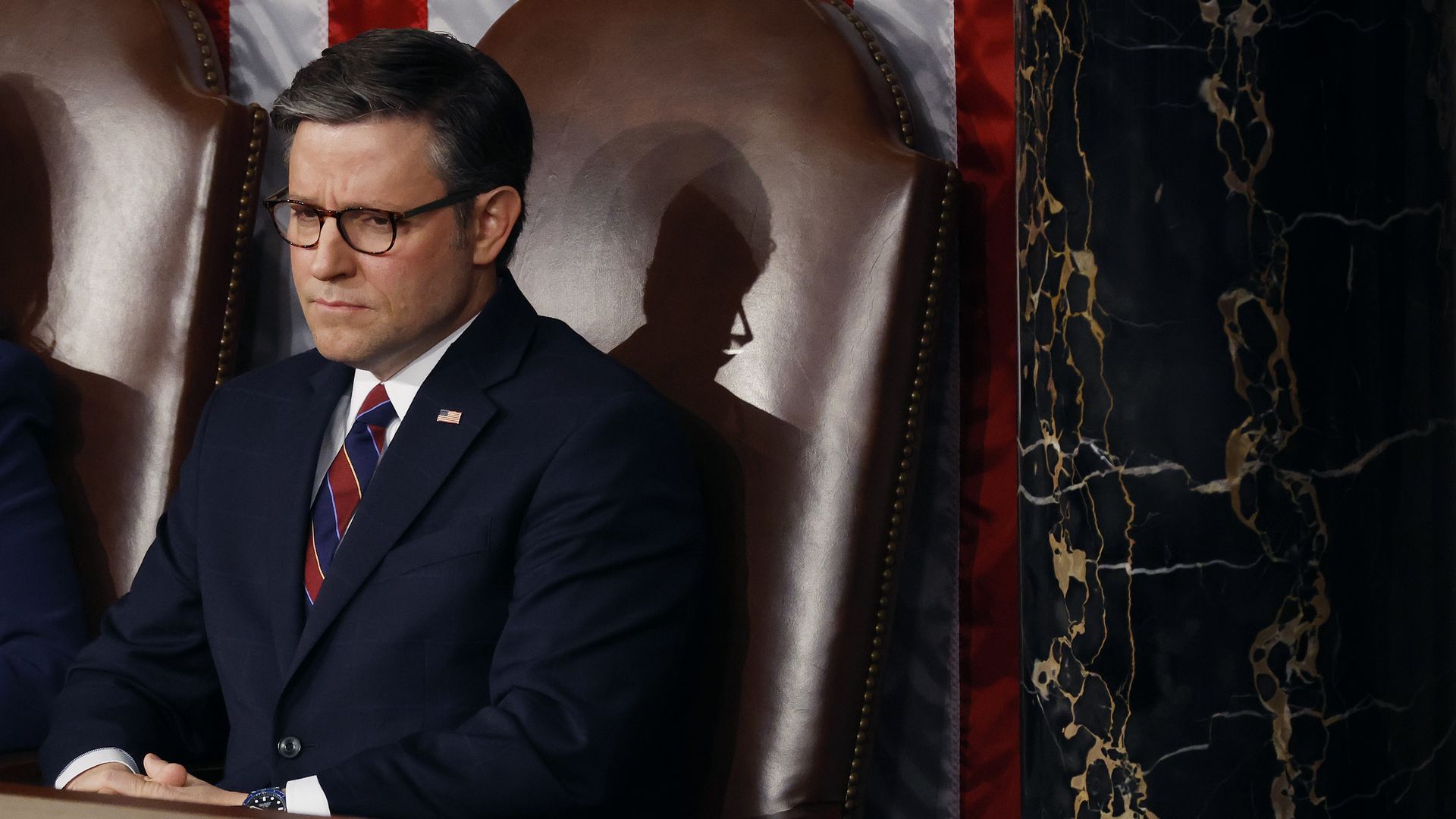 House Speaker Mike Johnson, wearing a dark blue suit, white shirt, red and blue striped tie and glasses, sitting in a large brown leather chair in the House chamber.