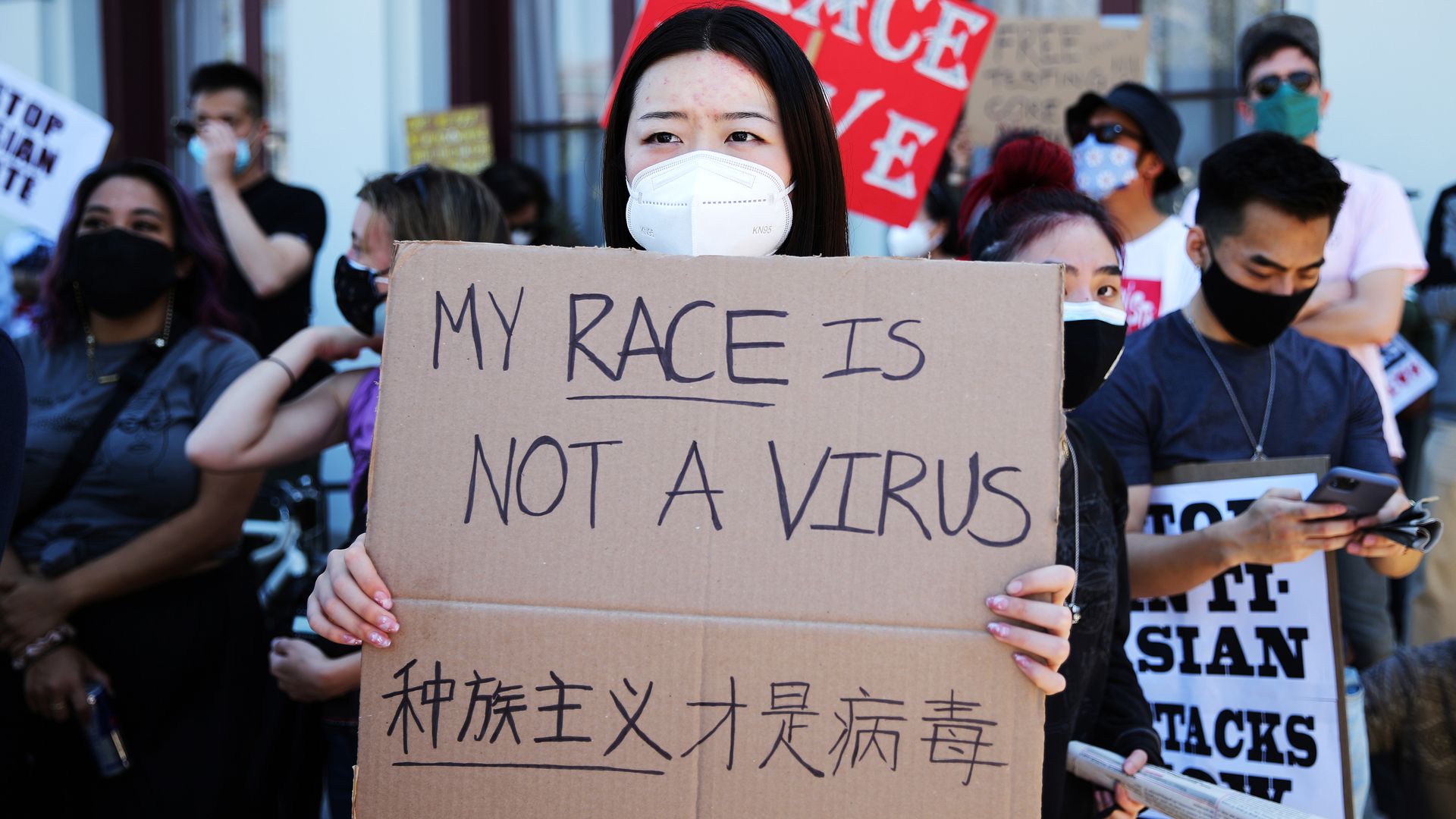 Photo of a masked Asian protester holding a cardboard sign that says "My race is not a virus" in English and Chinese