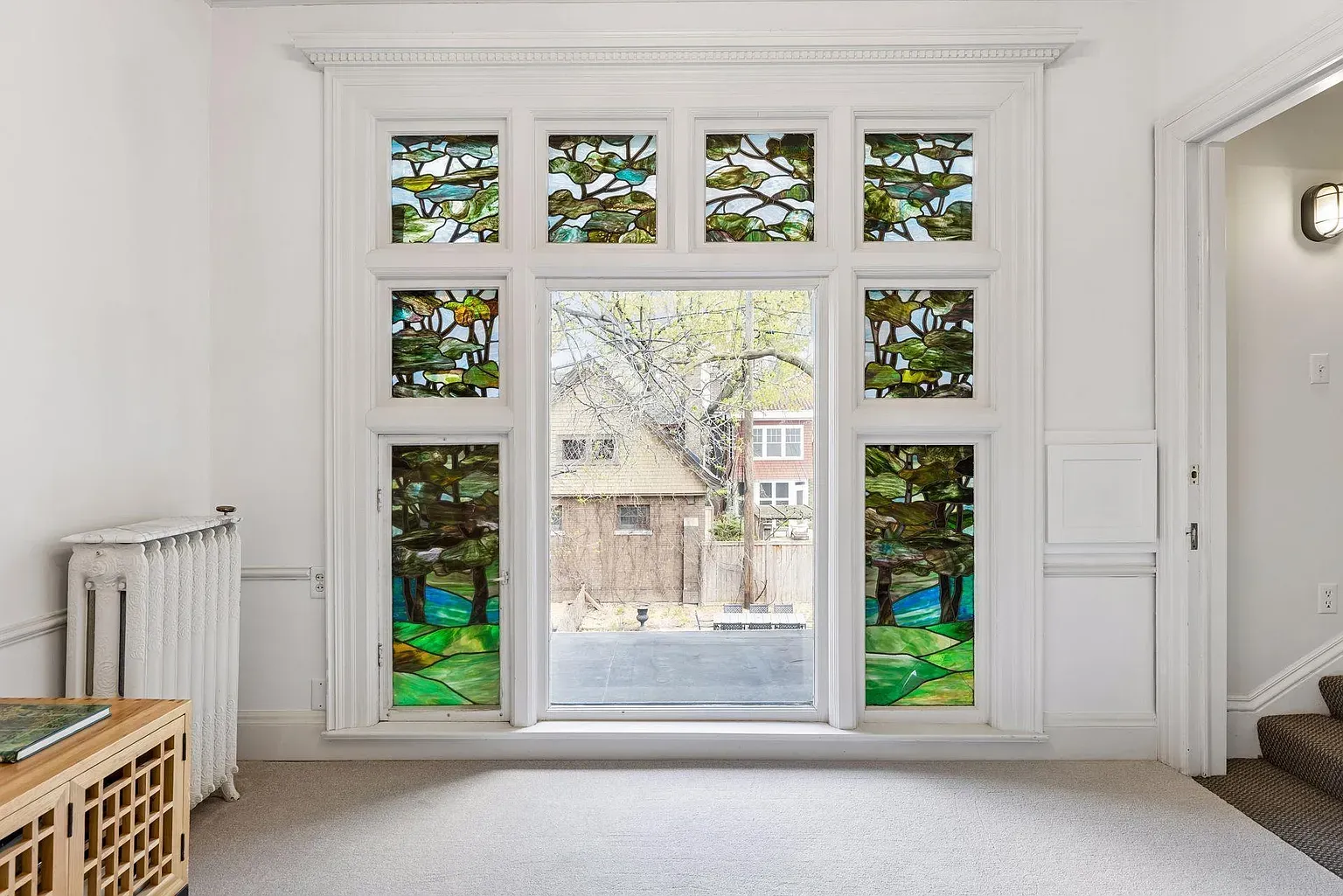 White room with a large central window framed by decorative trim, flanked by stained-glass panels depicting green trees and hills. Outside, a yard and neighboring houses. Left: radiator and a wooden bench; right: stairs.