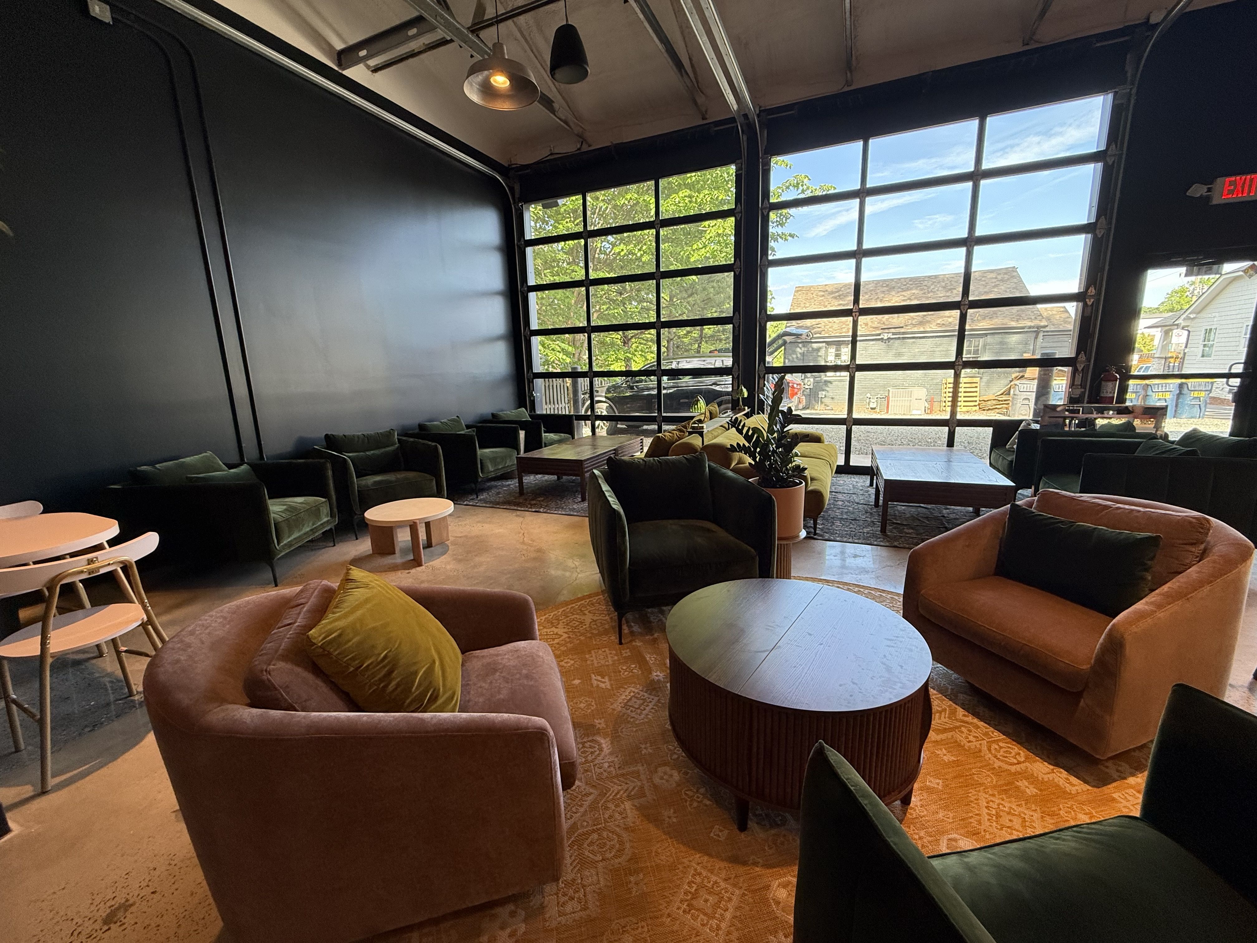 Cozy modern lounge with dark walls and a wall of glass windows; olive-green and blush seating, round wooden coffee tables, and an orange patterned rug, with daylight and city houses visible outside.