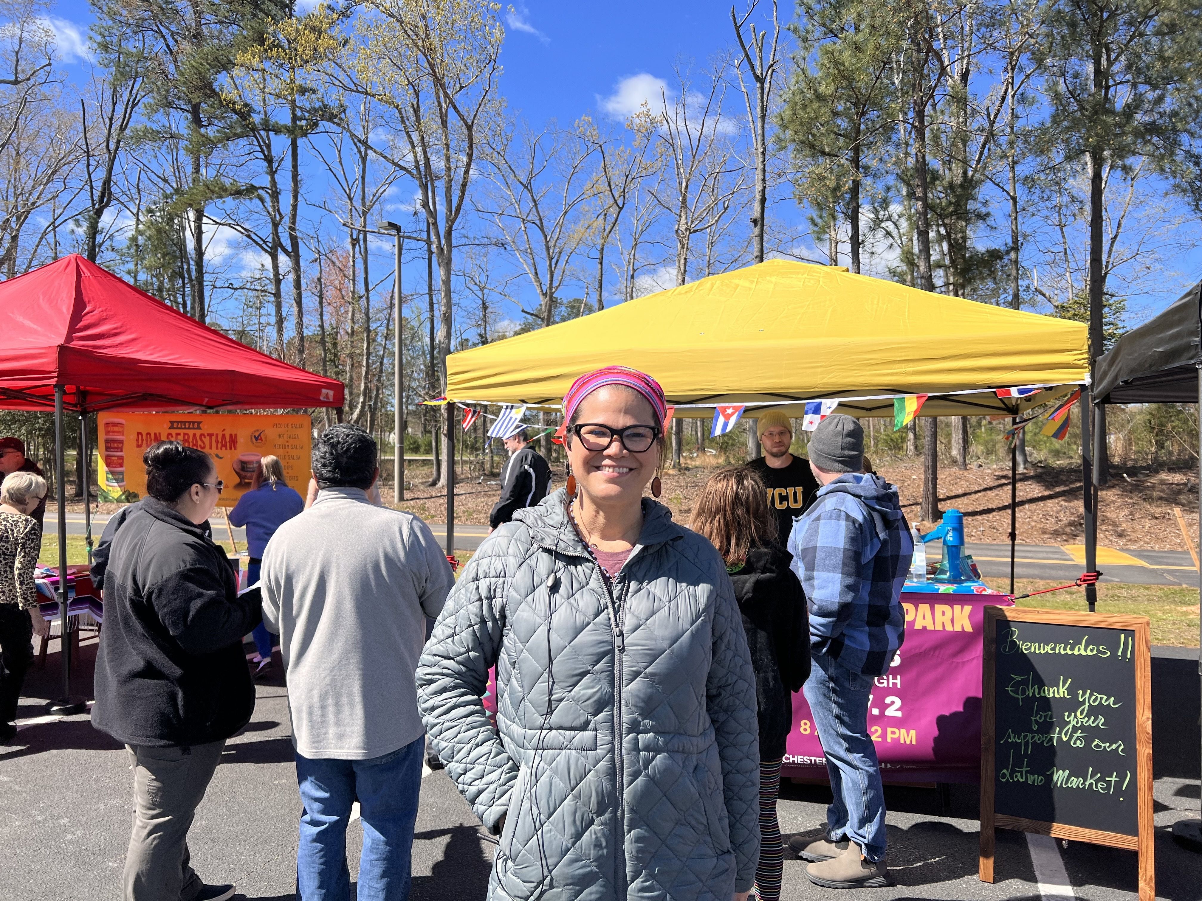 A woman smiling at the camera with people and red and yellow tents. One has a sign for Don Sebastian, another has flags hanging from them and a sign that says "Bienvenidos!! Thank you for your support to our Latino market!"