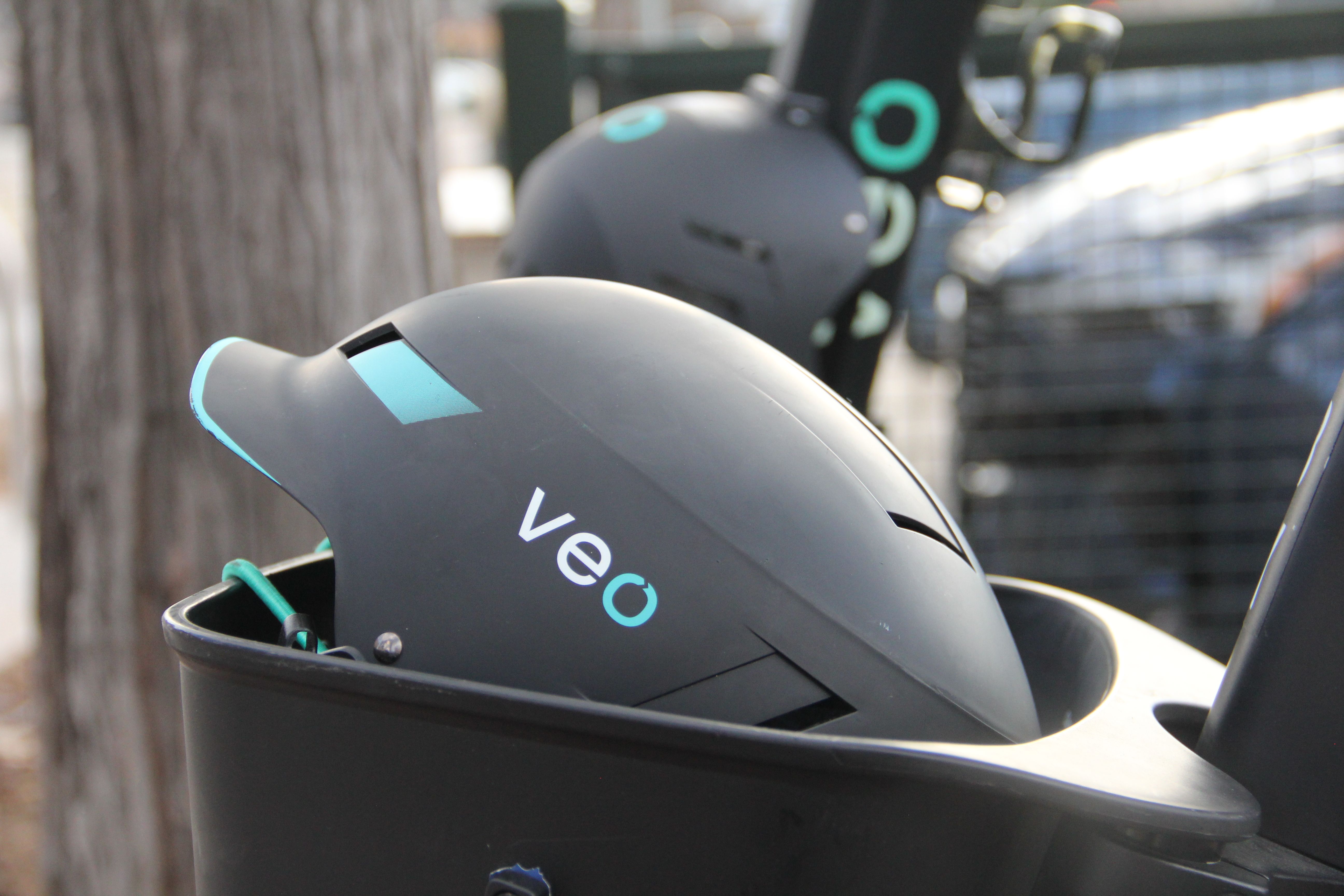 Black helmet with blue accents and the word "veo" in white, placed in a black container attached to a vehicle, with a blurred tree and background behind it.