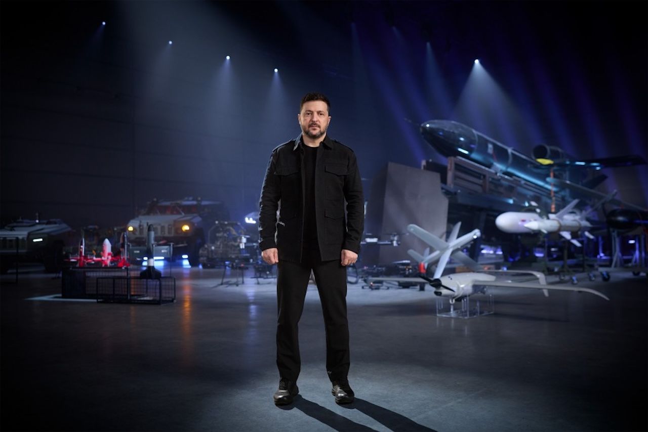 Volodymyr Zelensky stands in a dim, blue-lit aircraft hangar filled with exhibits, including jets and missiles, behind him as spotlights shine from above.