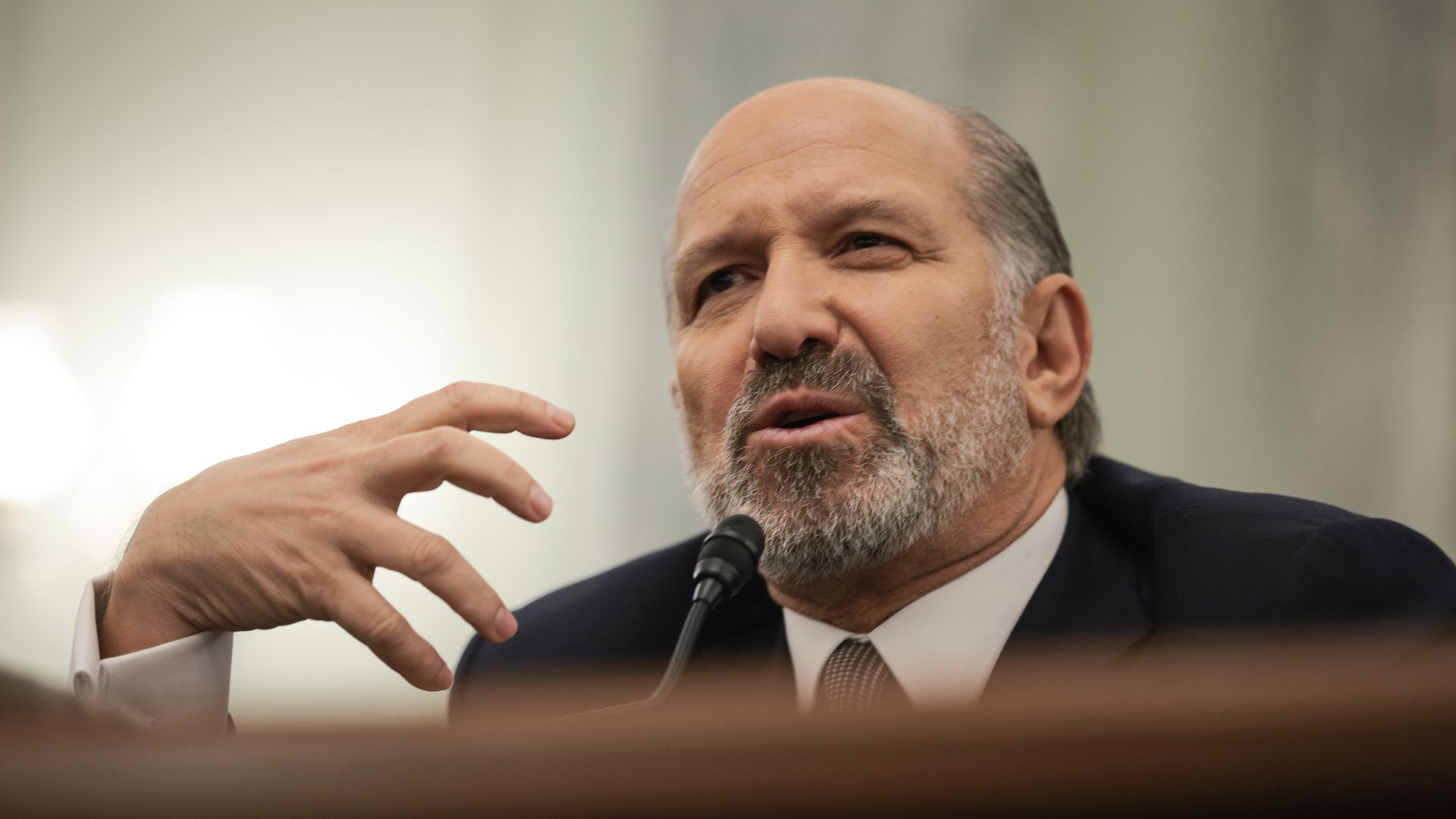 A mature man speaking in a suit from the shoulders up, gesturing with one hand. 