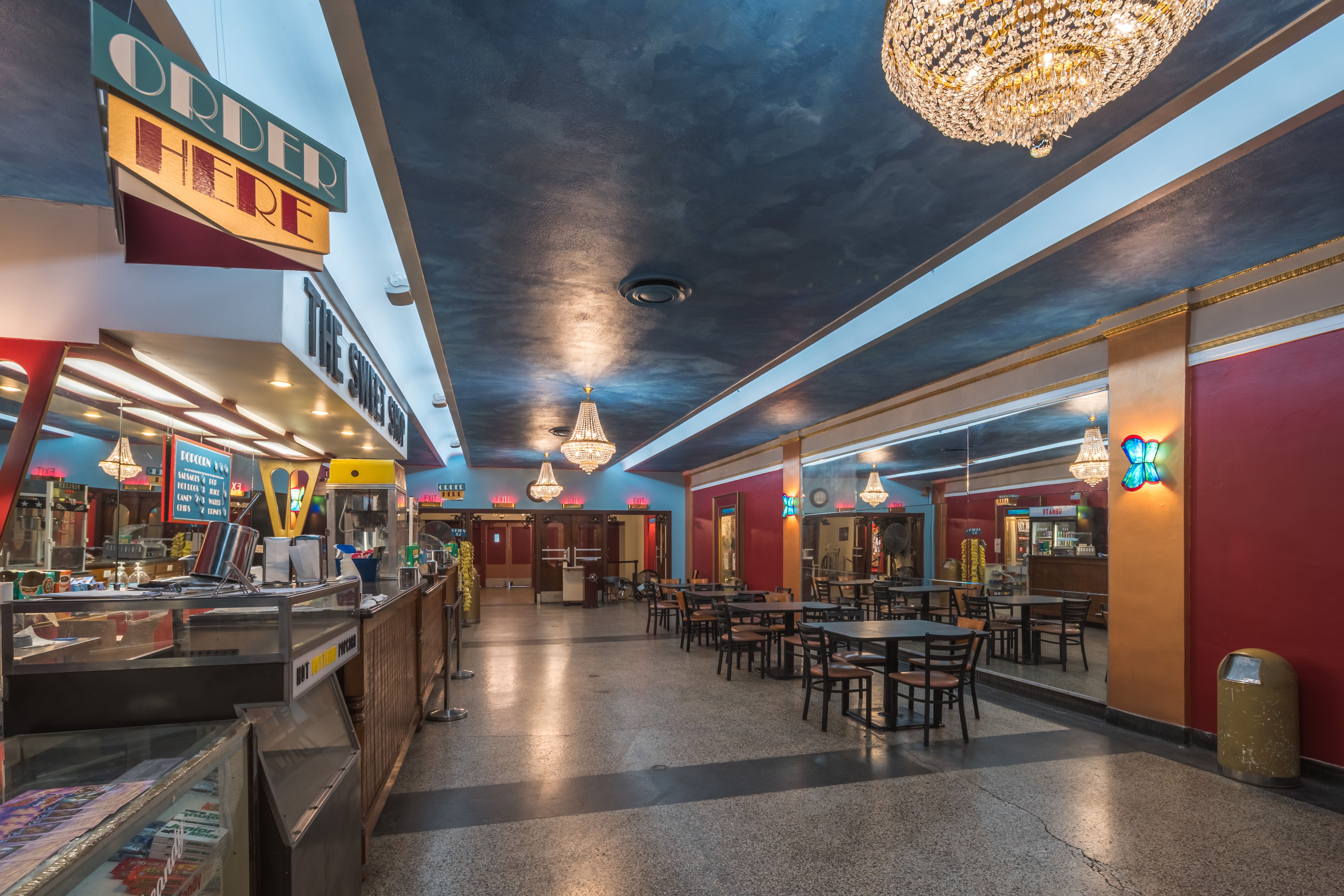 Interior of a retro concession stand: blue ceiling with crystal chandeliers, glass display counters to the left, colorful signs, and rows of tables and chairs beneath warm lighting.