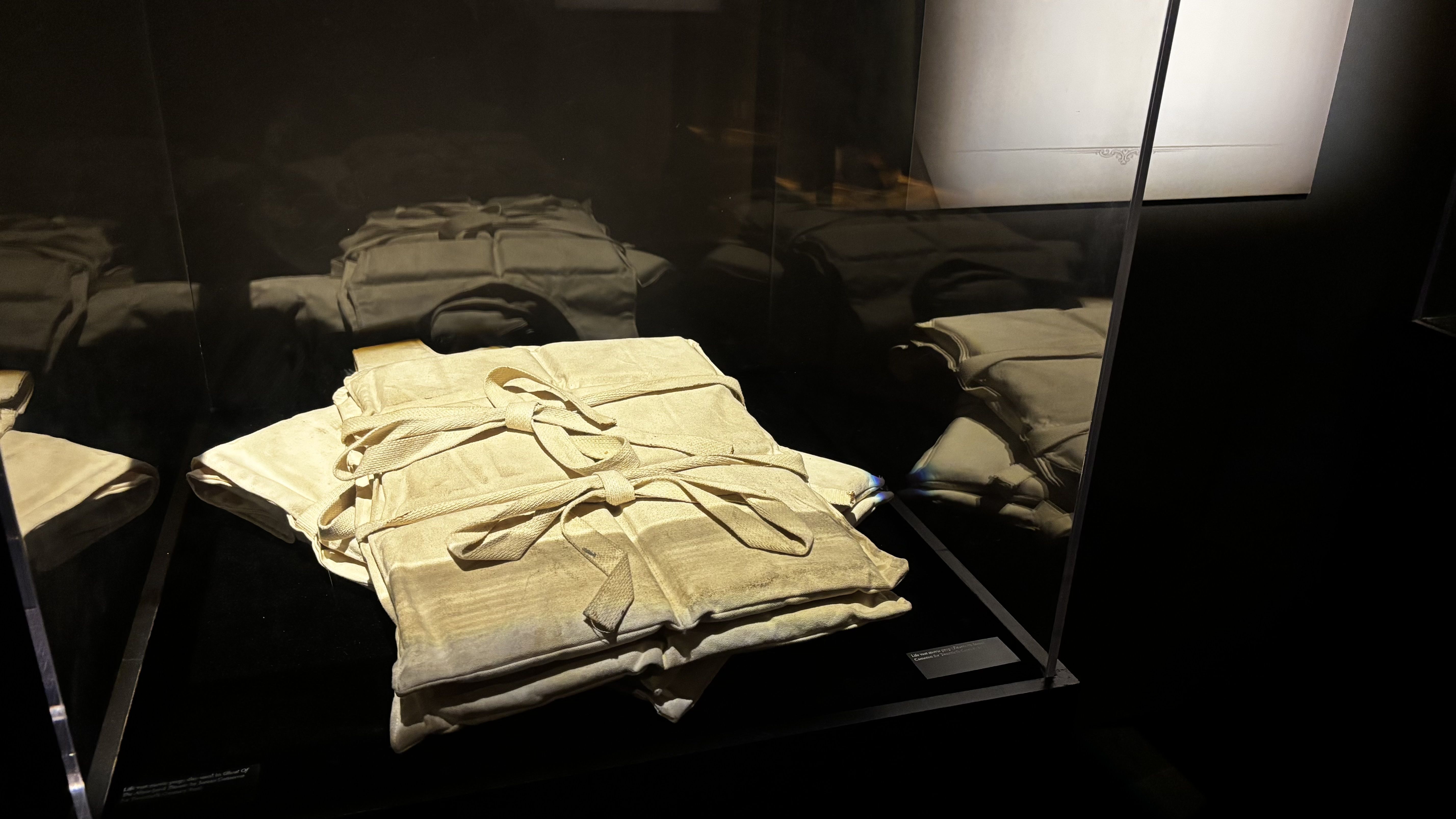 Beige straightjacket with straps tied on a black surface, displayed in a glass case with dark reflections and a lighted label on the right.