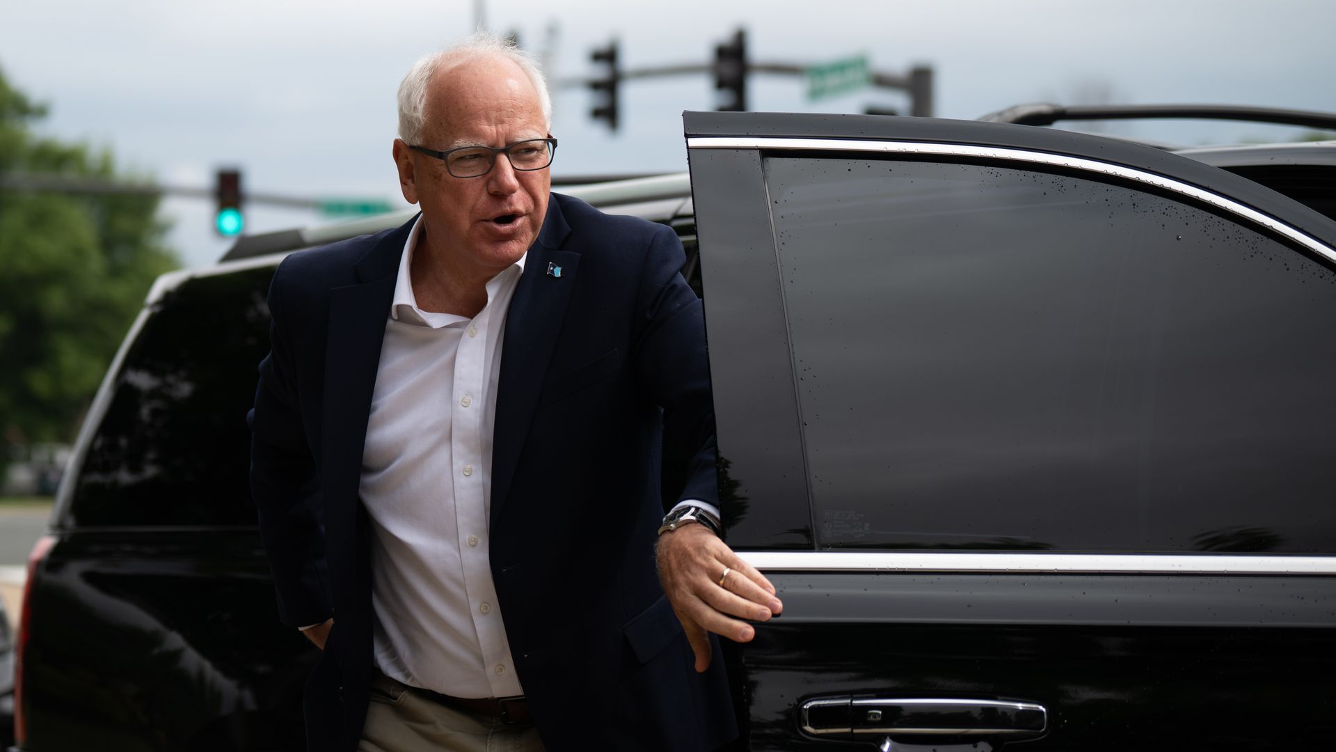 Tim Walz, wearing a blue suit and no tie, steps out a black car.