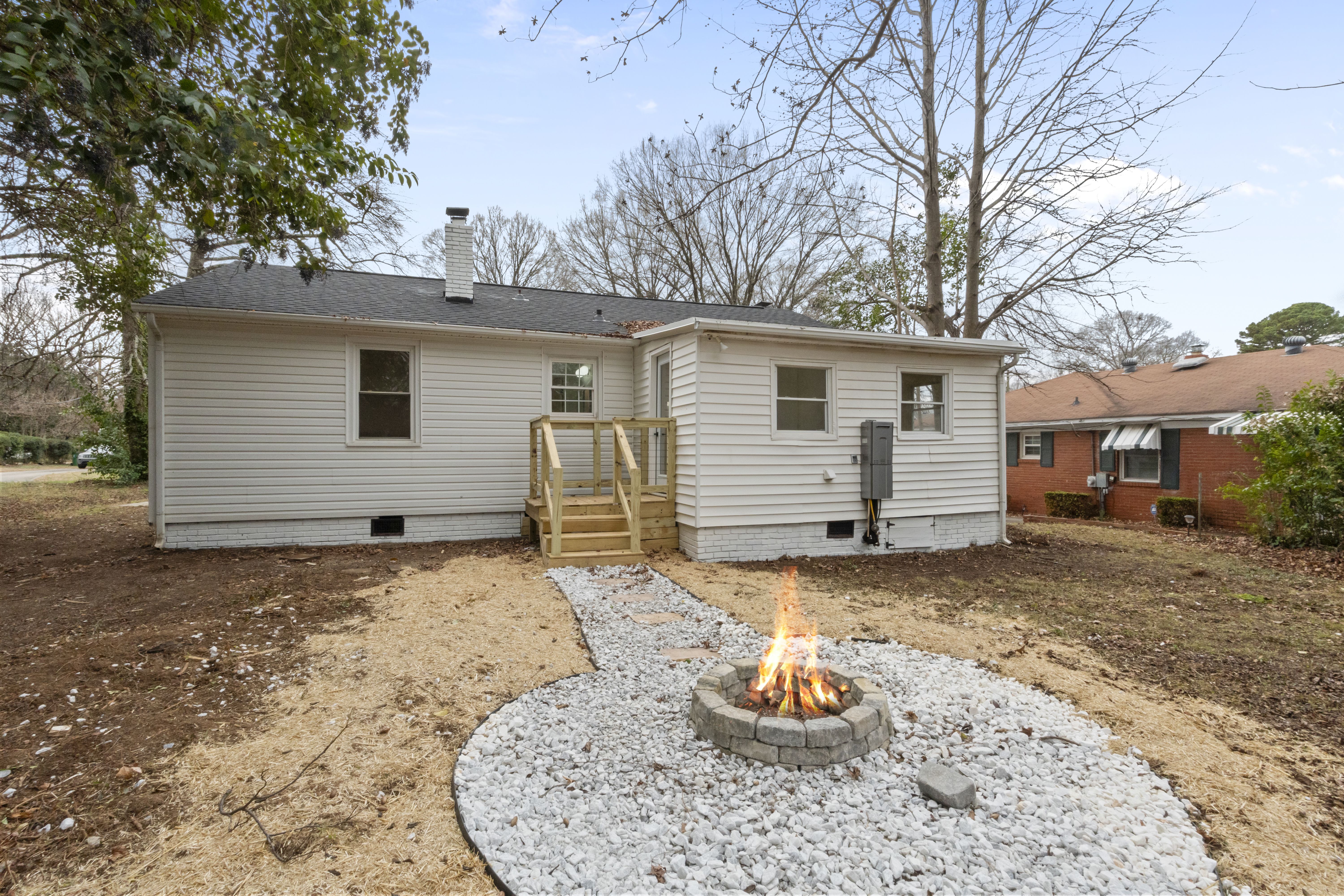 Back of a white single-story house with small wooden steps leading to a door. In the yard, a lit fire pit surrounded by white gravel and a stone path. Leafless trees and neighboring brick house visible.