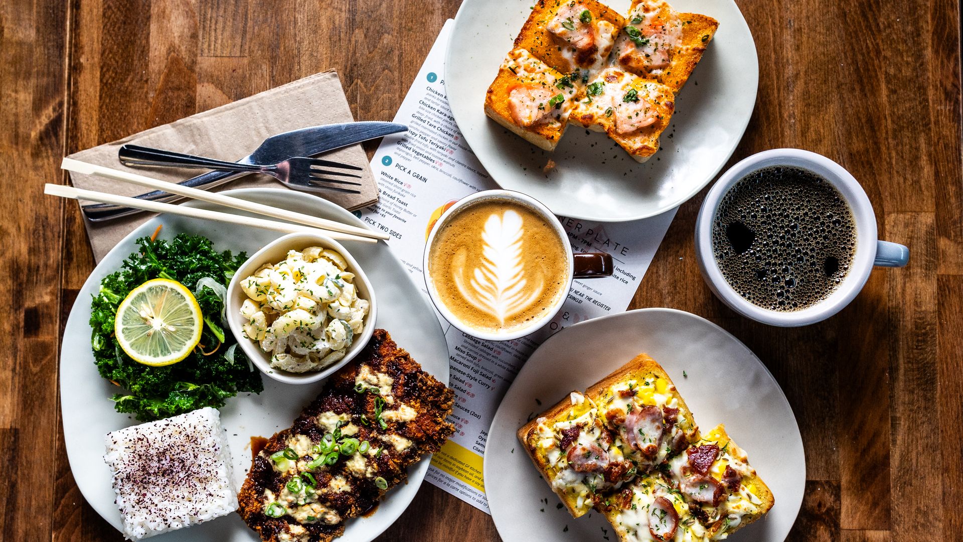 Top-down view of a brunch spread on a wooden table: cheesy herb toasts, kale with lemon, potato salad, a latte with leaf art, and a cup of black coffee with silverware nearby.