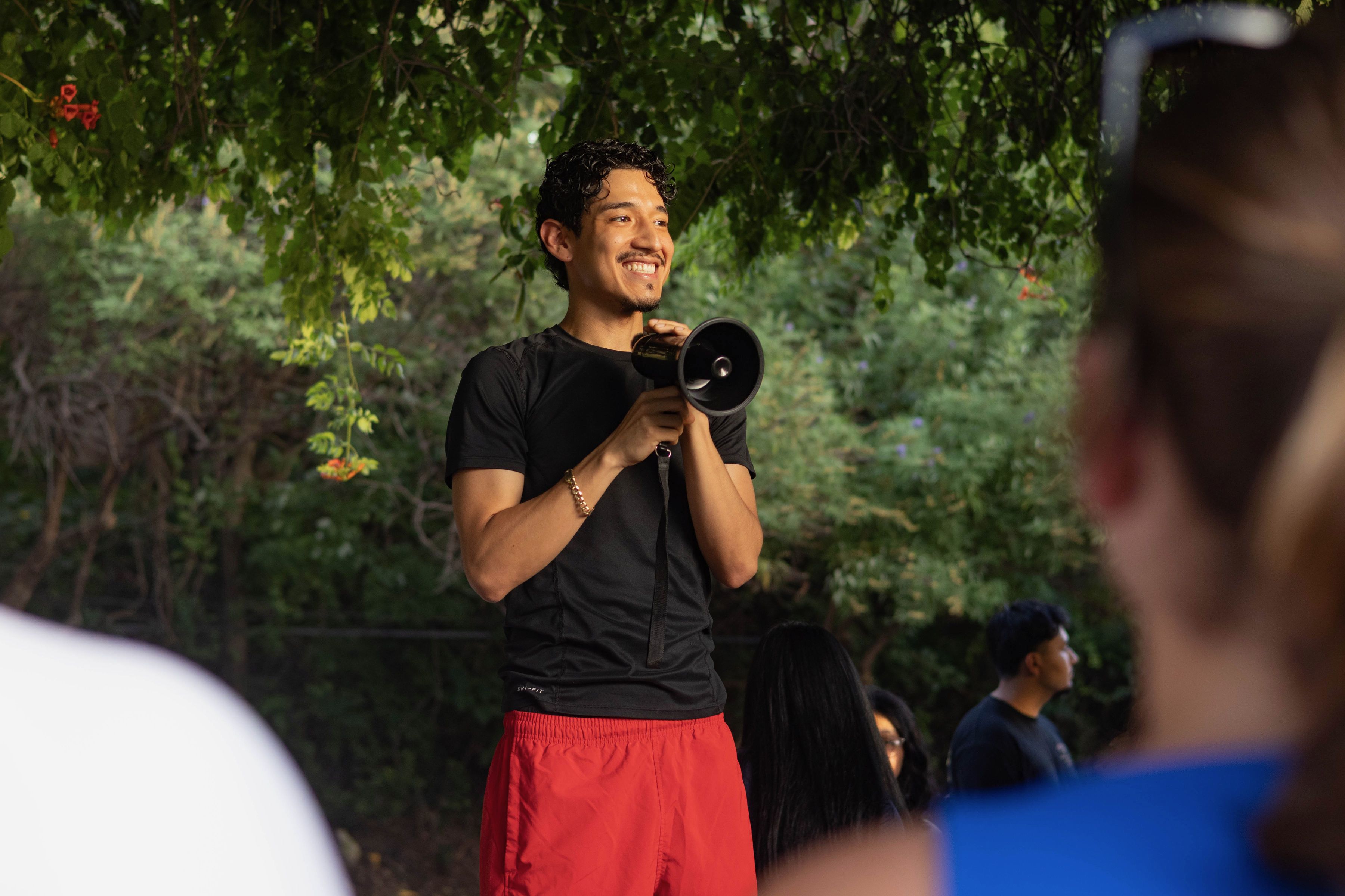 Smiling man with dark curly hair wearing a black T-shirt and red shorts holds a megaphone outdoors, surrounded by green foliage; blurred people sit and stand in the background.