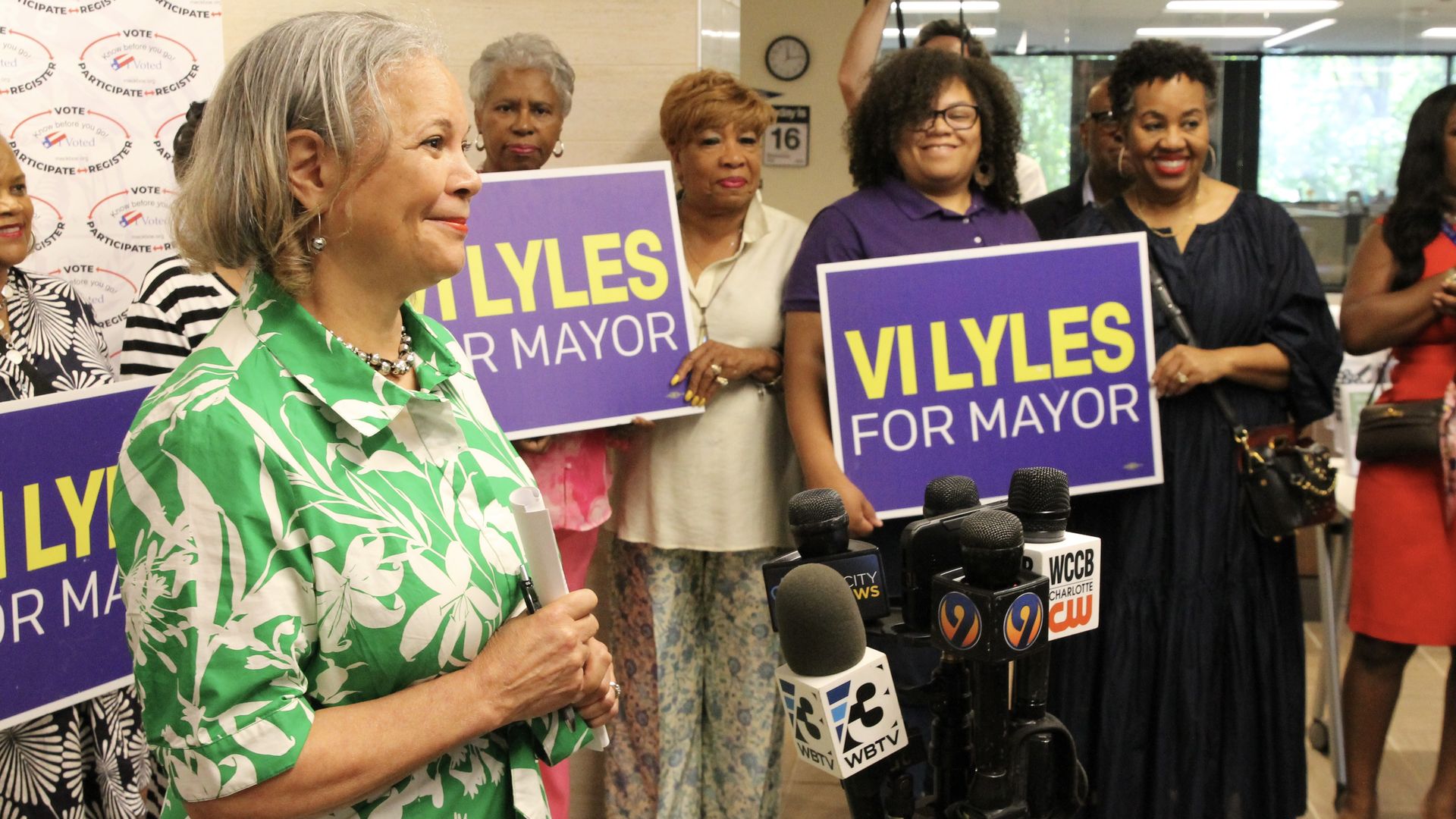 Woman in green and white floral dress speaking at a press event with supporters holding purple and yellow "VI LYLES FOR MAYOR" signs behind her, with microphones in front.