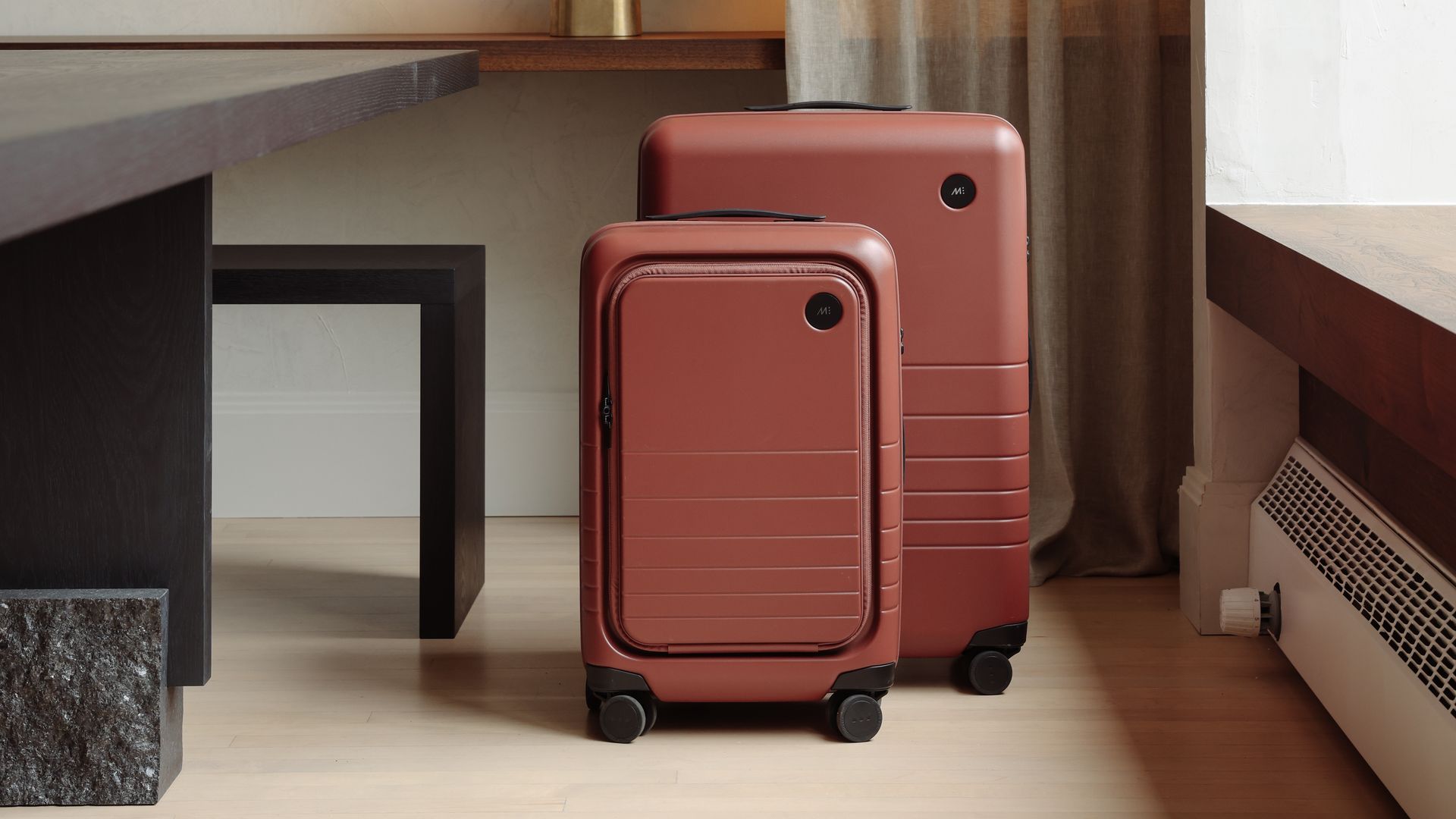Two pieces of luggage, both a dark tan color, sit between a window and a table.