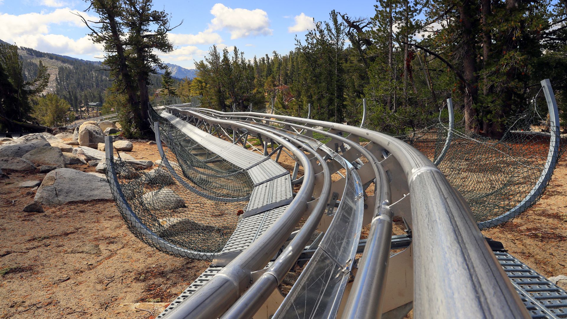 Winding silver roller-coaster tracks curve along a rocky, pine-covered mountainside, fenced by chain-link barriers. Rocks, trees, and distant peaks under a bright blue sky with clouds.