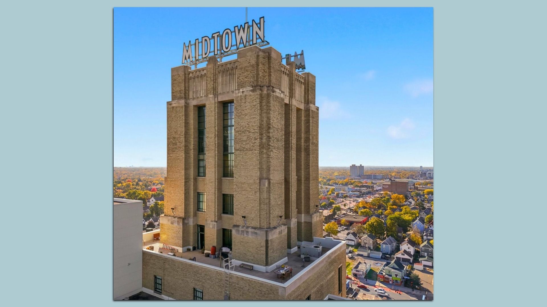 Tall beige brick building with large "MIDTOWN" sign on top, surrounded by cityscape with trees and houses under clear blue sky on a sunny day.