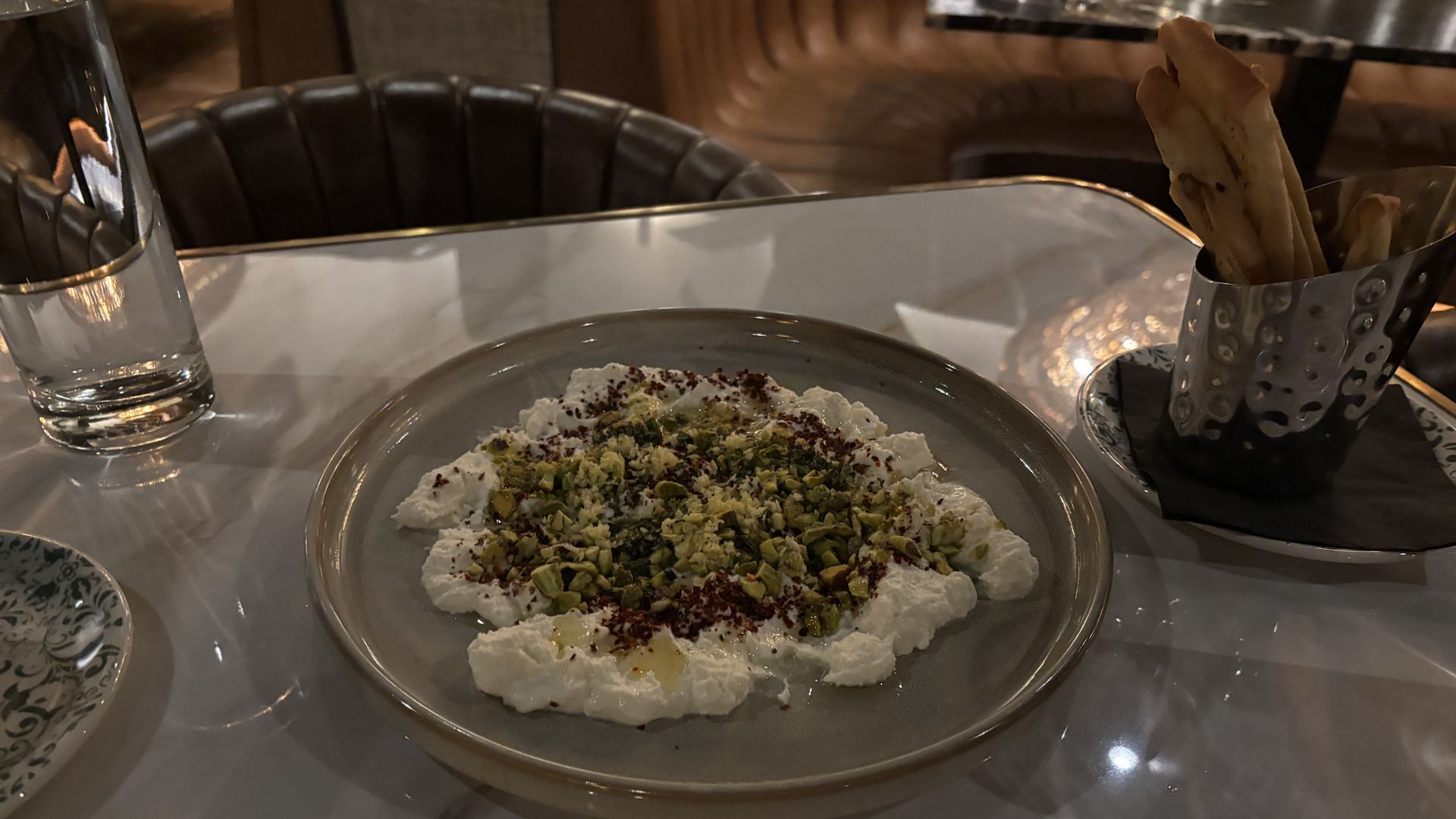 Plate with spreadable white cheese topped with chopped pistachios and spices on a white marble table, next to a glass of water and a metal cup with breadsticks.