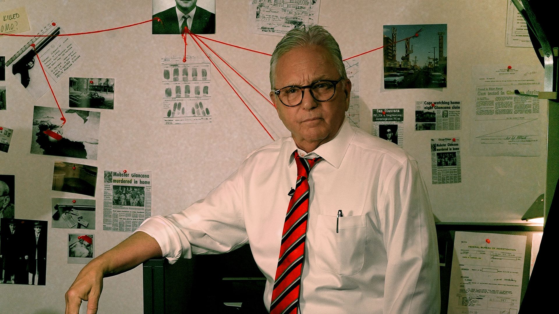 Photo of a man posing in front of a evidence board with photos and strings attaching clues. 