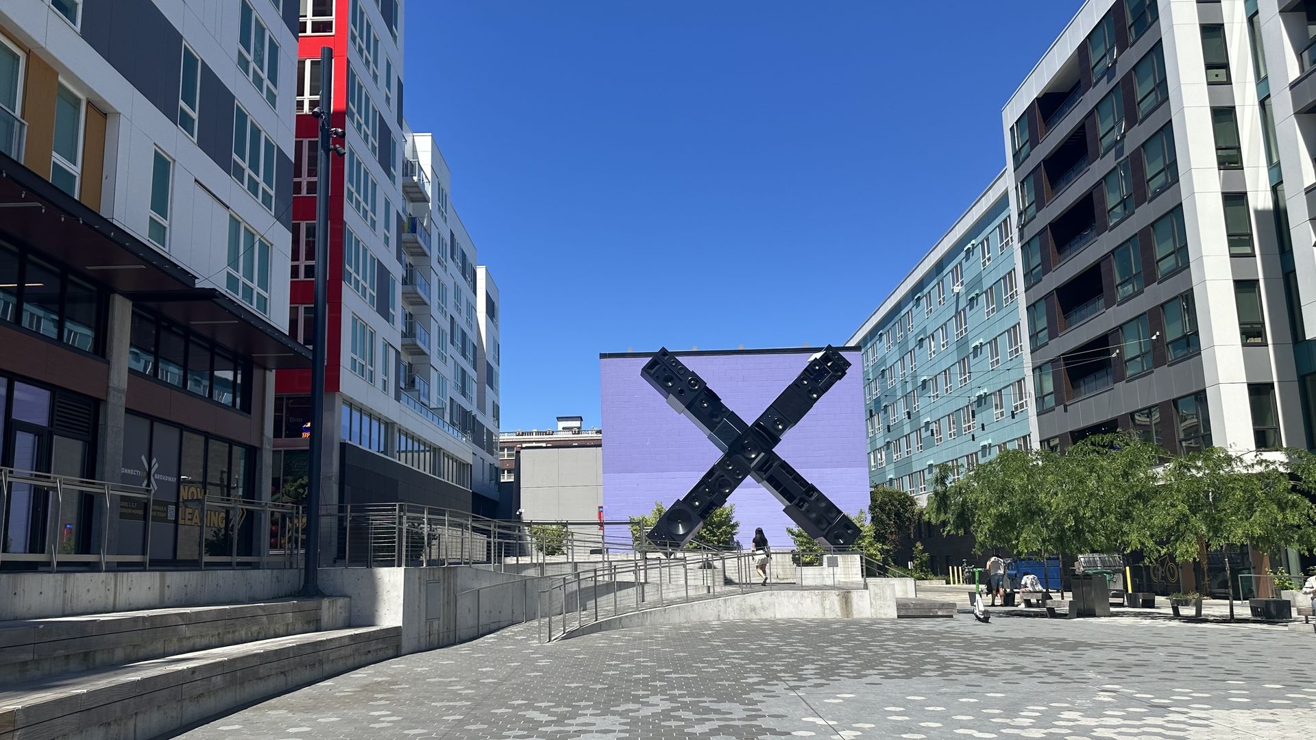 Wide urban plaza between modern white-gray buildings on both sides. A large black X sculpture sits before a purple wall on hexagonal tiled pavement; a few people walk under a clear blue sky.