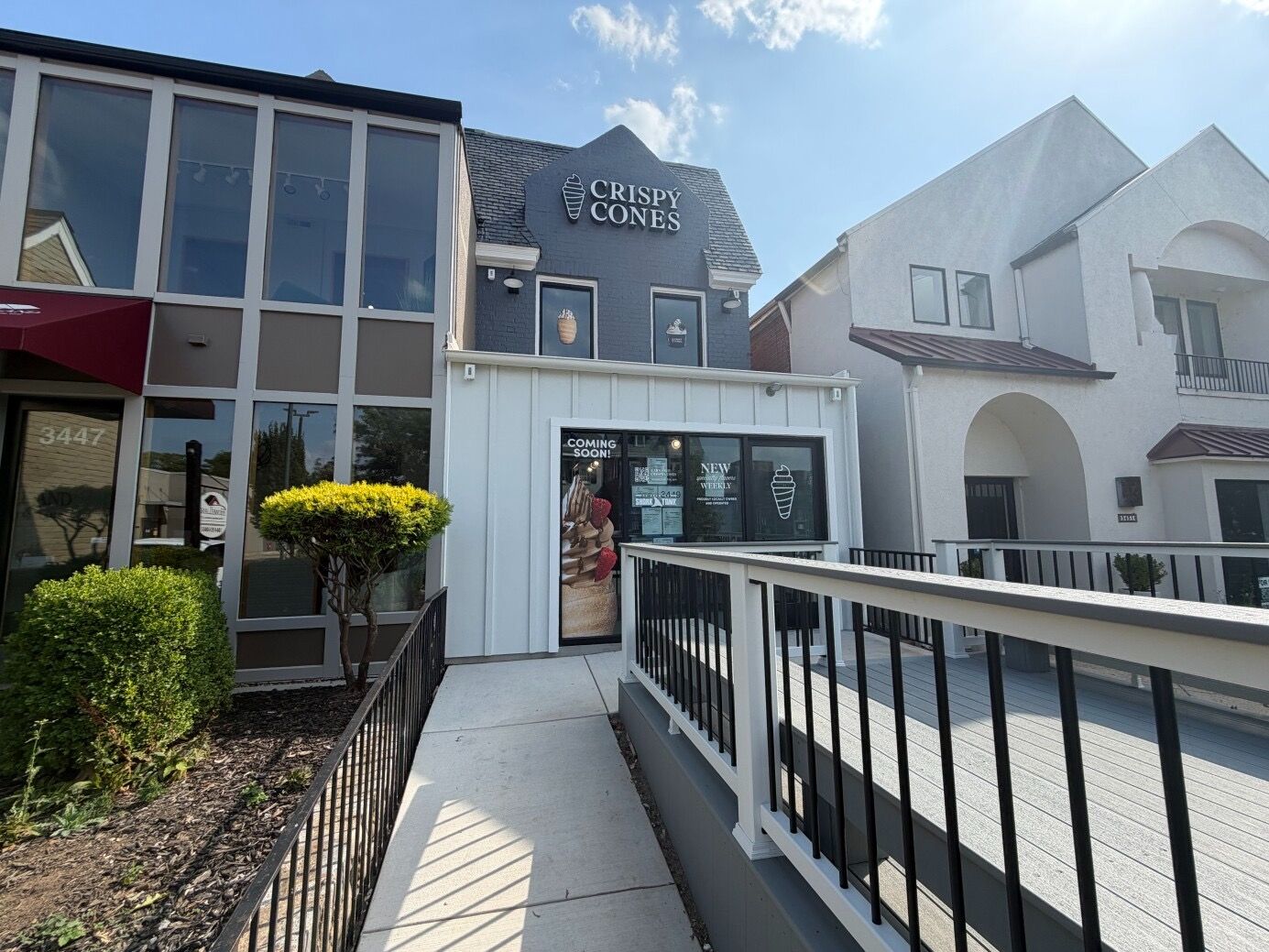 Street view of a row of shops, featuring the gray building with glass storefronts and the ice cream shop CRISPY CONES, a door poster, white railing, and a blue sky.