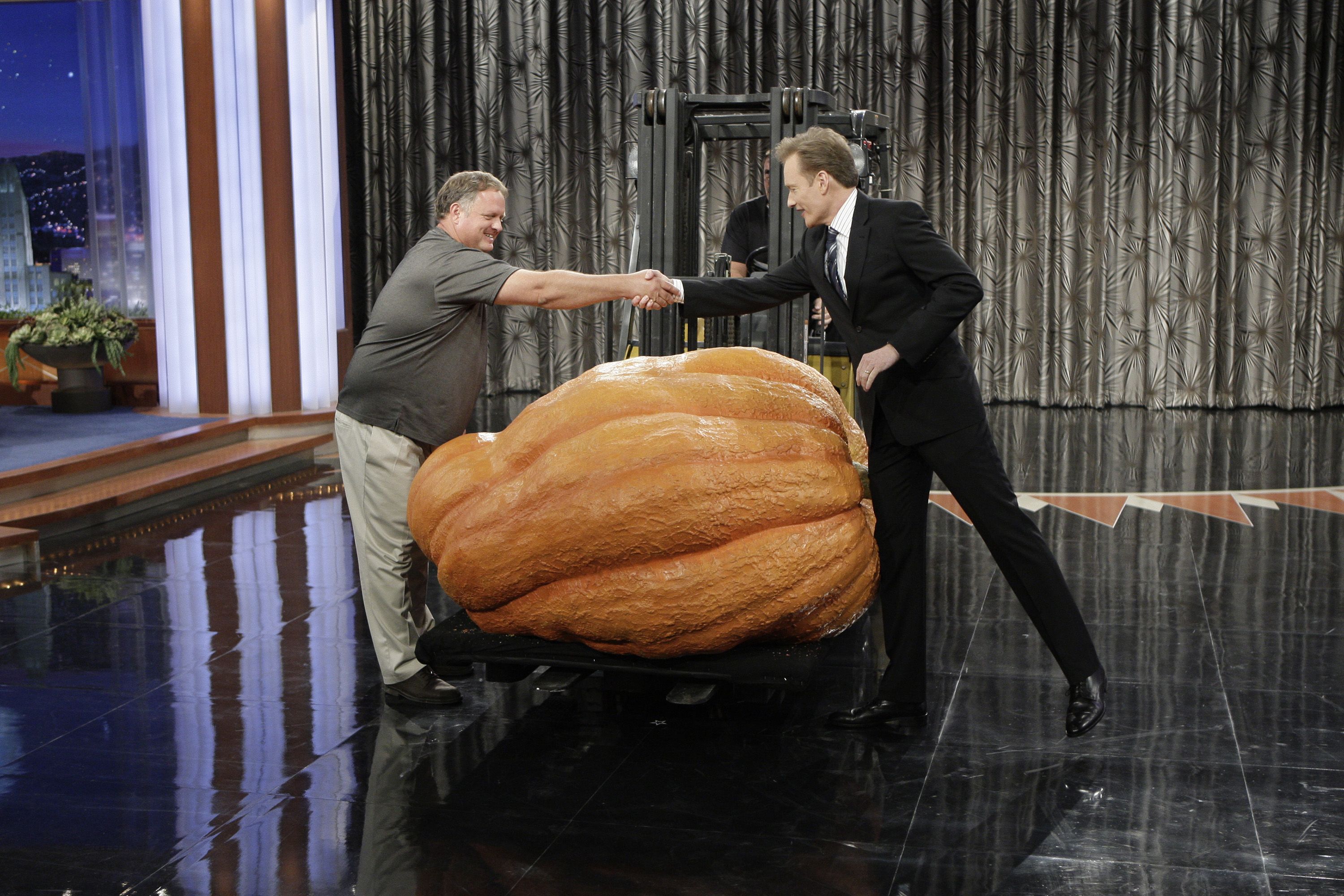 Two men on a talk-show stage shake hands over a giant orange pumpkin sculpture on a wheeled platform; one in a gray shirt, the other in a black suit, against a metallic curtain backdrop.