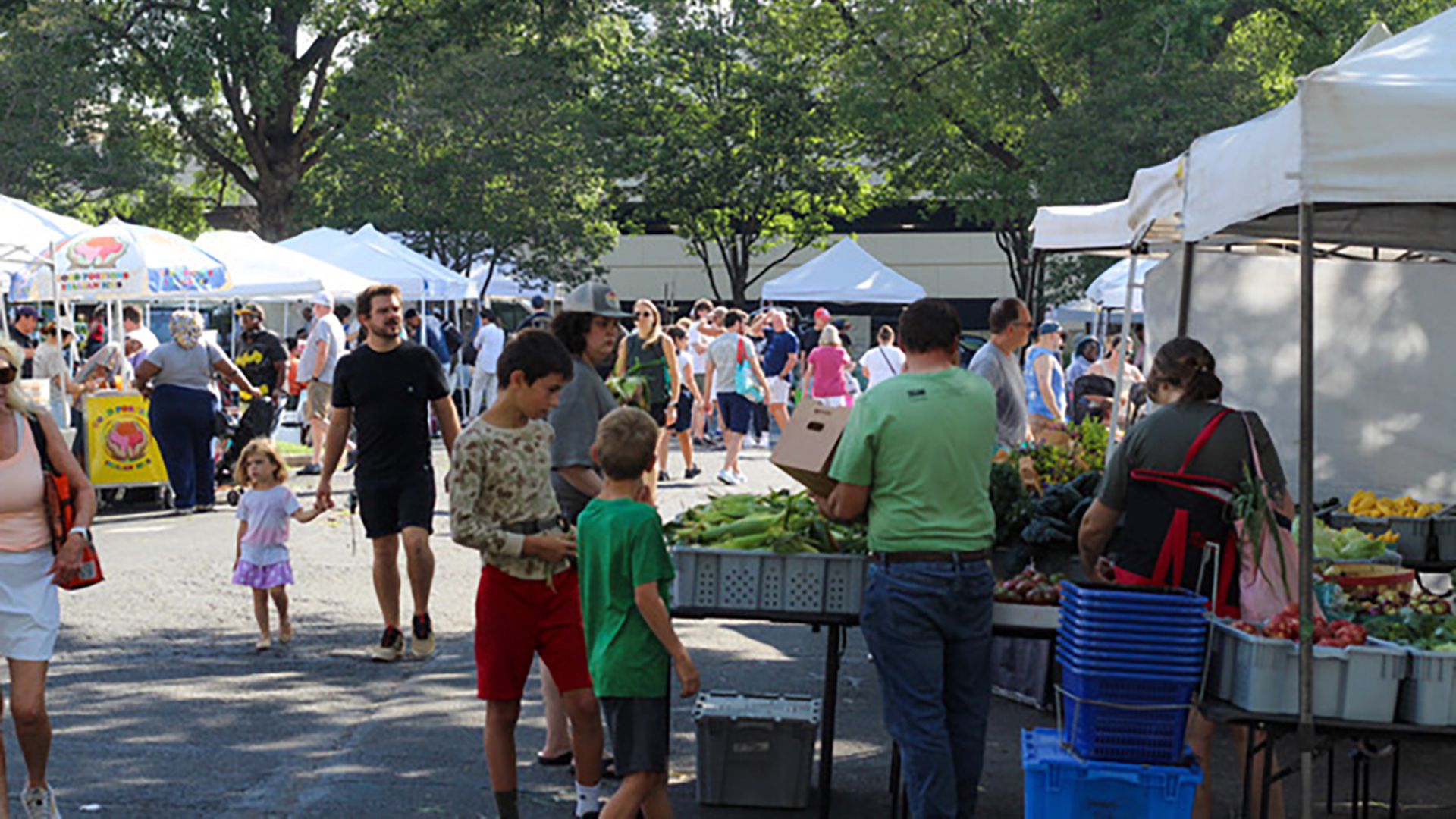 Outdoor farmers market on a sunny day with white canopies, leafy trees, and busy shoppers. A man in a green shirt browses fresh vegetables while kids and vendors mingle near crates of produce.