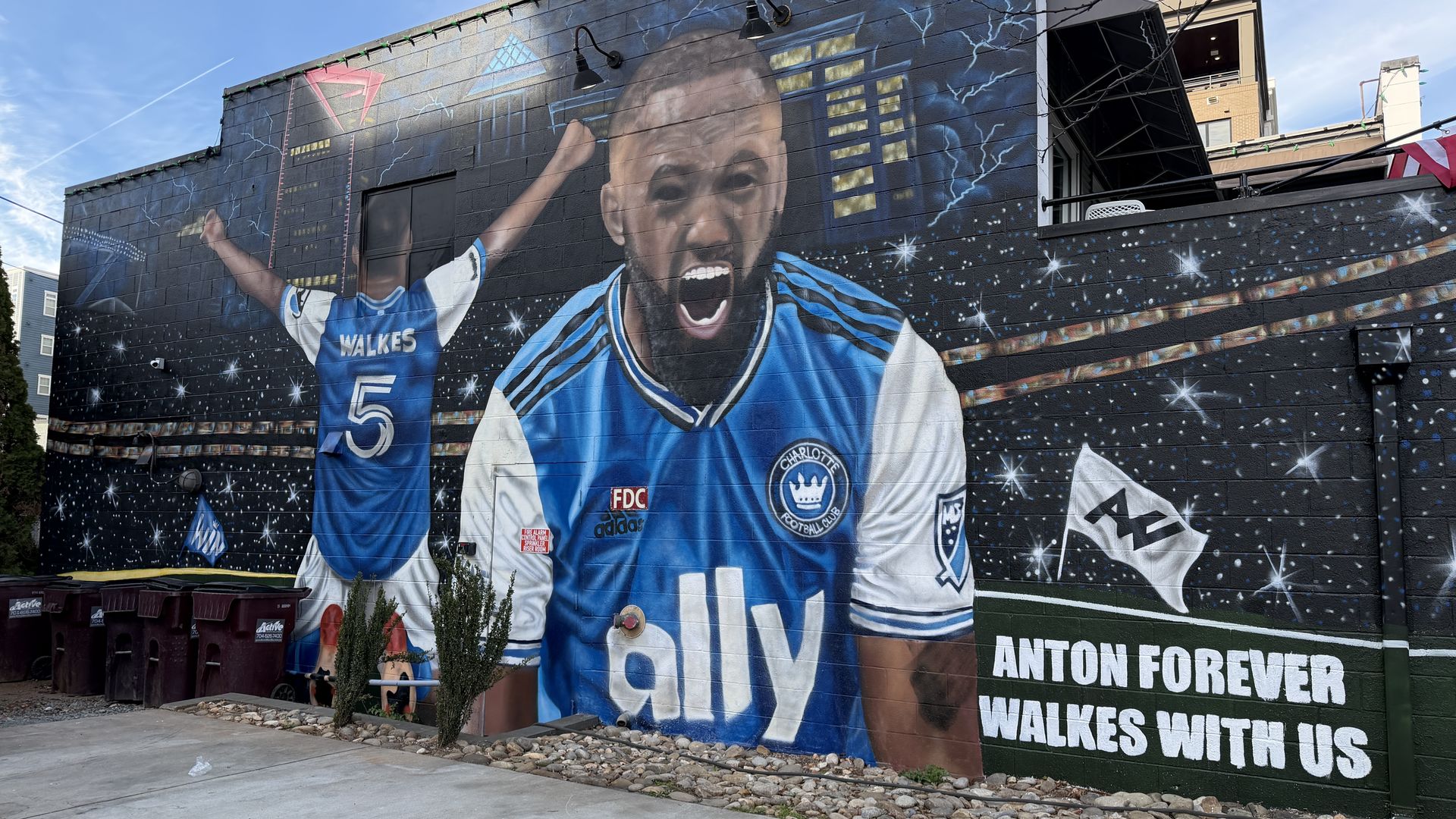 Mural of soccer player Anton Walkes in blue and white Charlotte FC jersey, celebrating with arms raised and shouting, cityscape and starry sky background, text: "Anton forever Walkes with us"