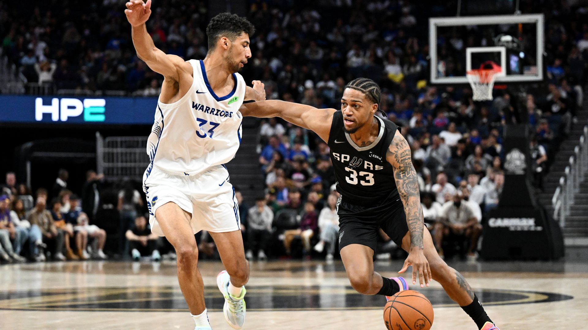 Two basketball players battle for the ball: a Warriors player in white jersey #37 and a Spurs player in black #33 drive toward the basket on a crowded arena court, with the hoop and crowd in sight.