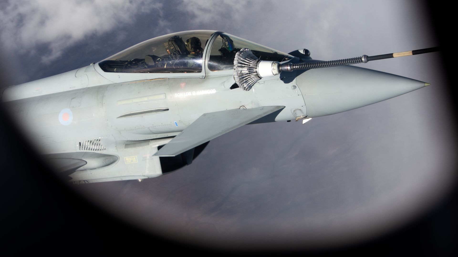 Gray military jet refueling in midair, with boom connected to its nose; pilot visible inside cockpit against cloudy blue sky background.