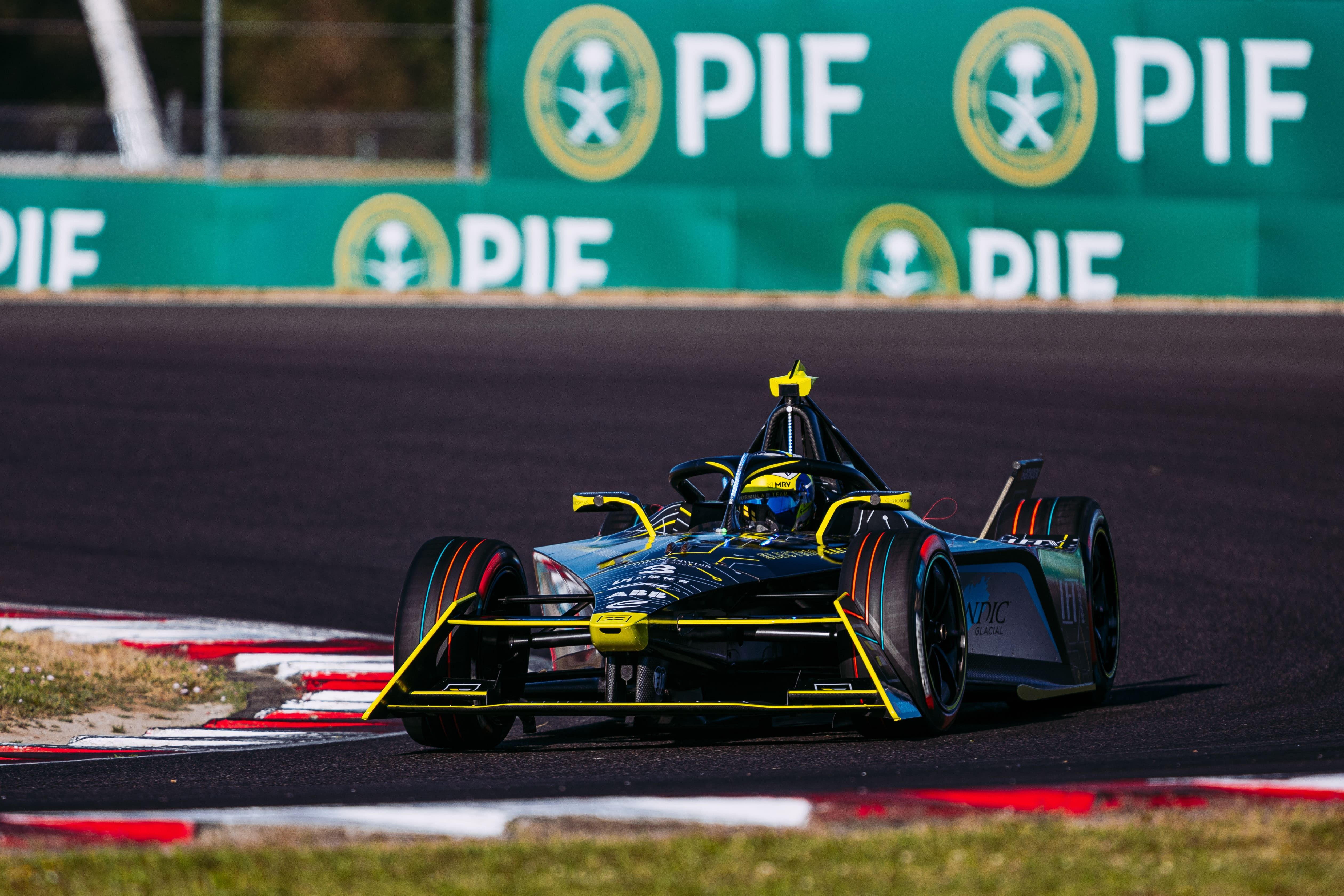 Black Formula E racecar with yellow accents on a racetrack corner, with green PIF sponsor banners in the background under clear weather conditions.