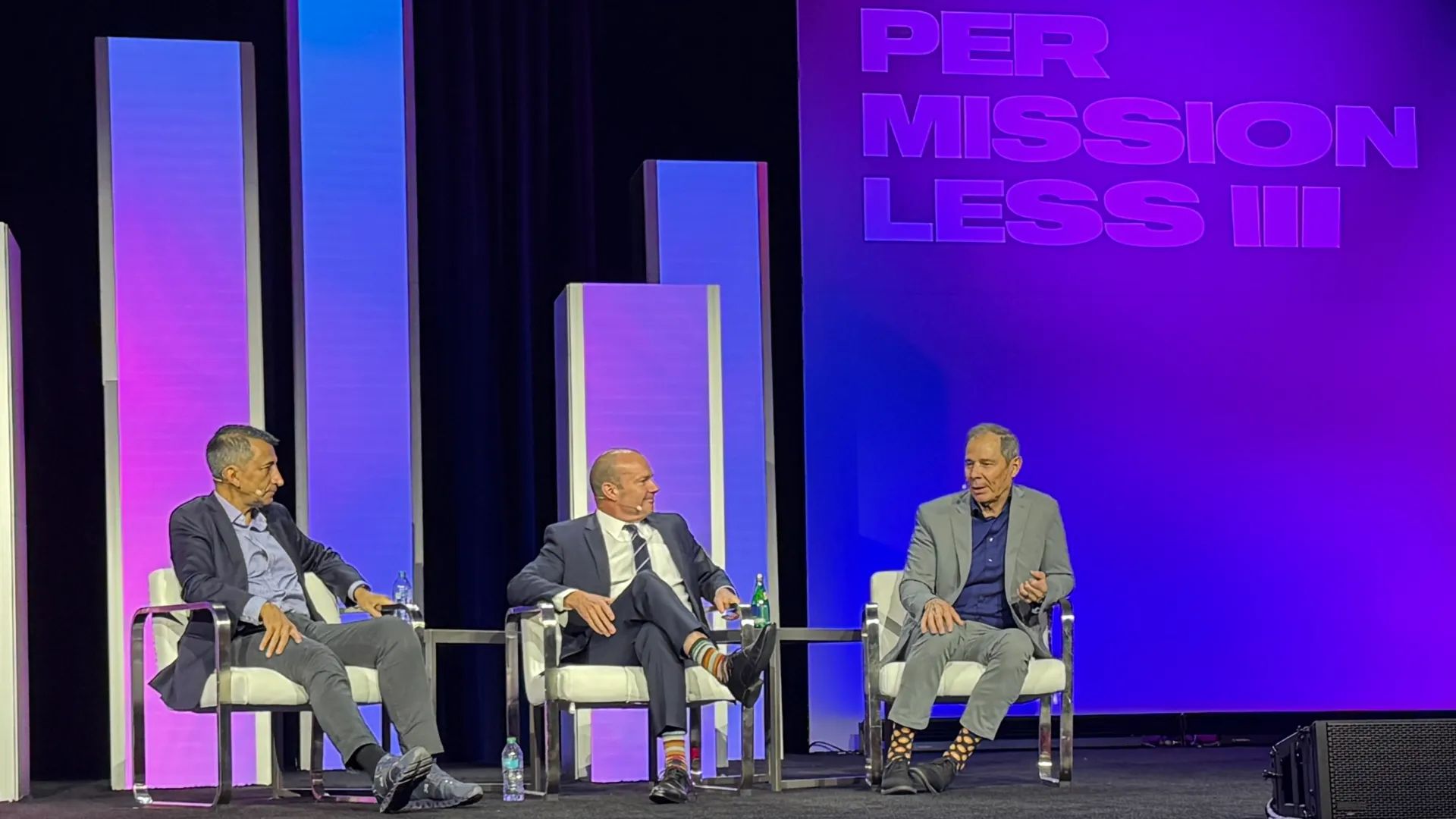 Three men sitting on a conference stage, talking. All in suit jackets. All distinguished looking. 