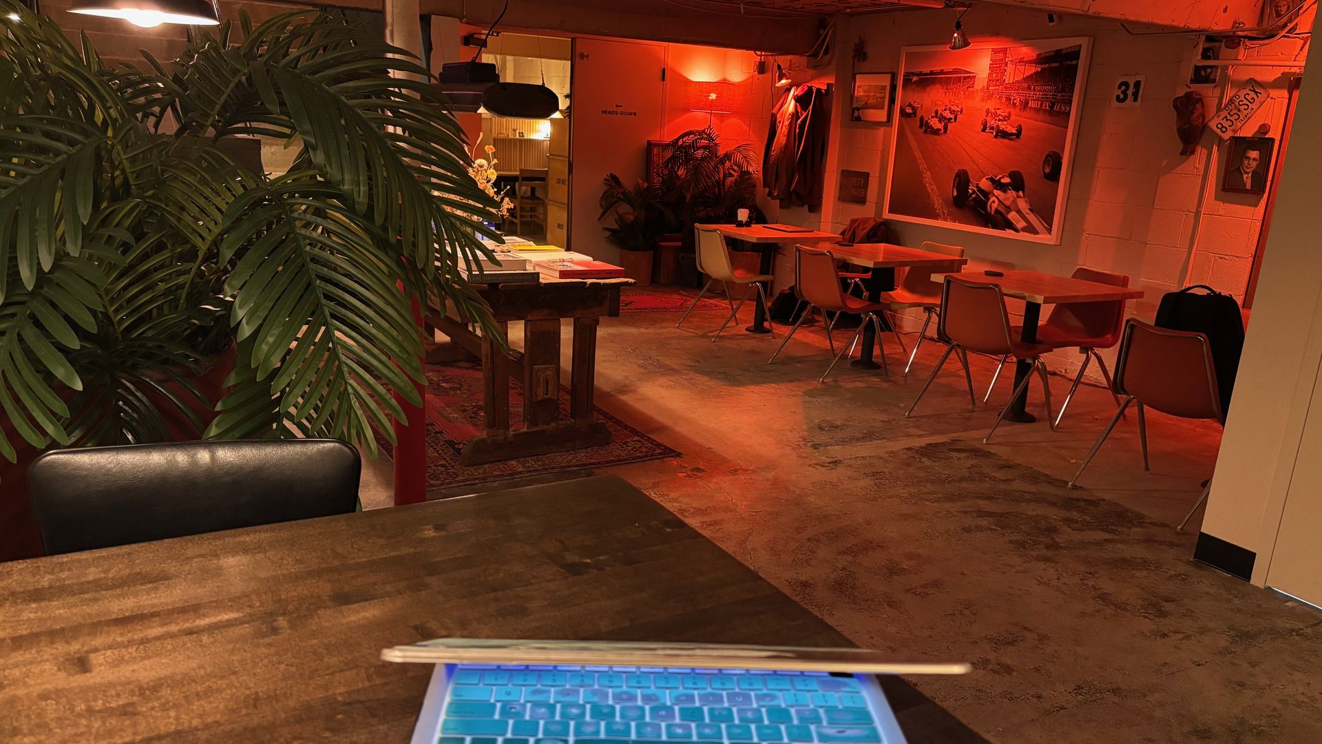 Red-lit cafe interior. A laptop on a dark wooden table in the foreground, with a large plant to the left. In the background, tables, beige chairs, and a wall mural of racing cars.