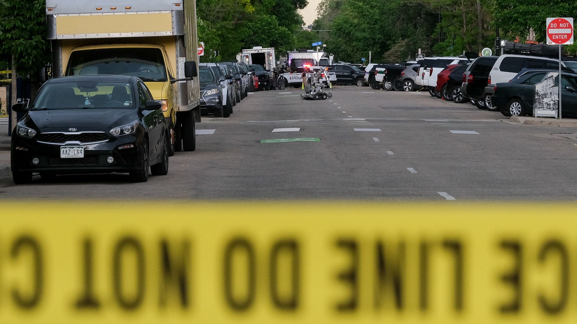City street lined with parked cars on both sides; a yellow box truck dominates the left foreground as police cars with flashing lights gather in the center and a robotic device sits on the road behind yellow tape.