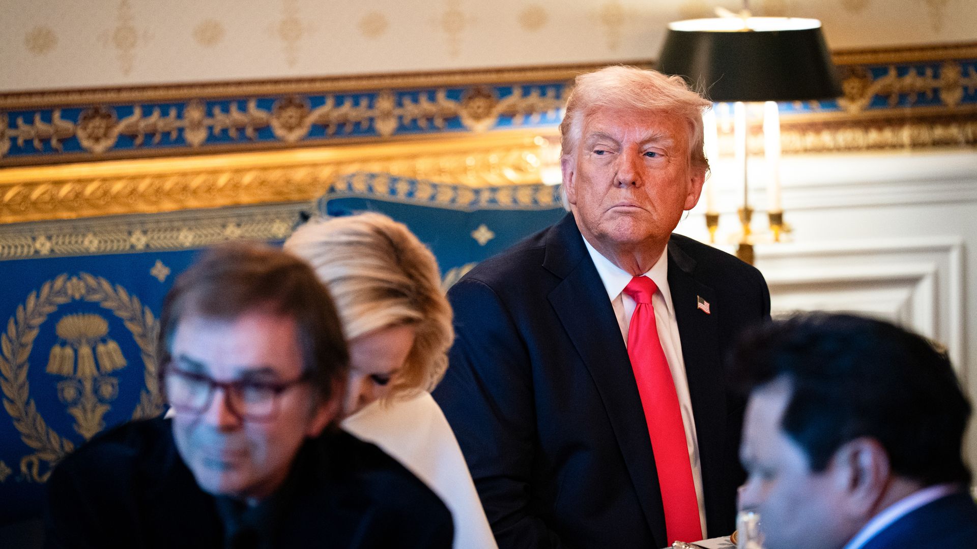 President Trump wearing a dark blue suit and a red tie, sitting at a table in an ornate blue-and-gold room with other people.