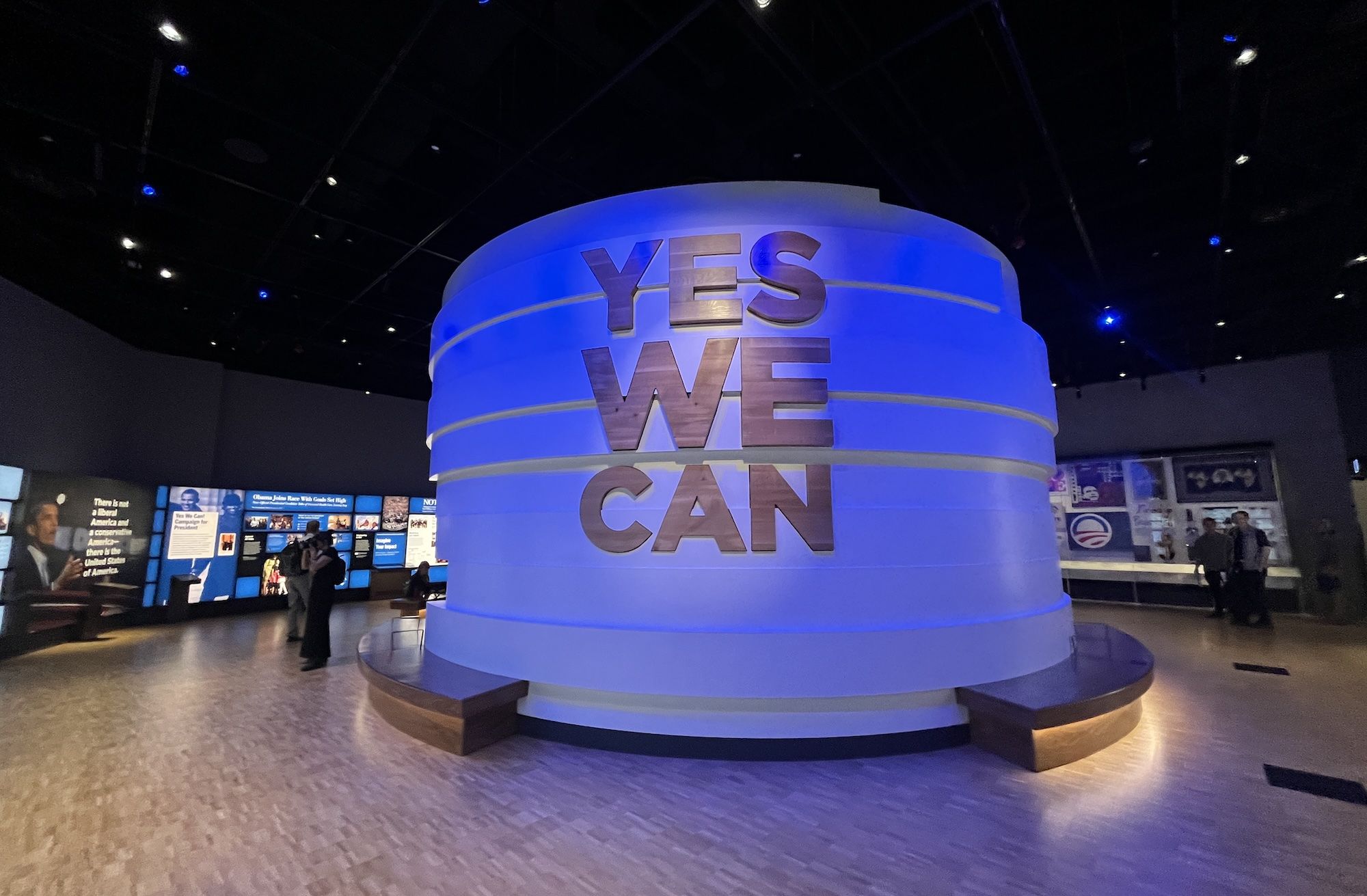Large blue cylindrical sculpture in a museum, with the silver letters YES WE CAN stacked around its tiers; blue lighting and display boards line the background.