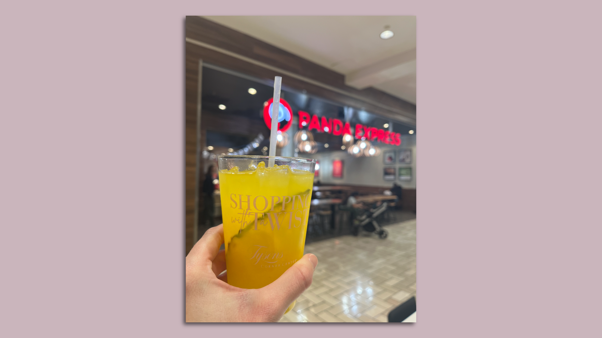 A hand holds a cocktail in front of a Panda Express at a mall. 