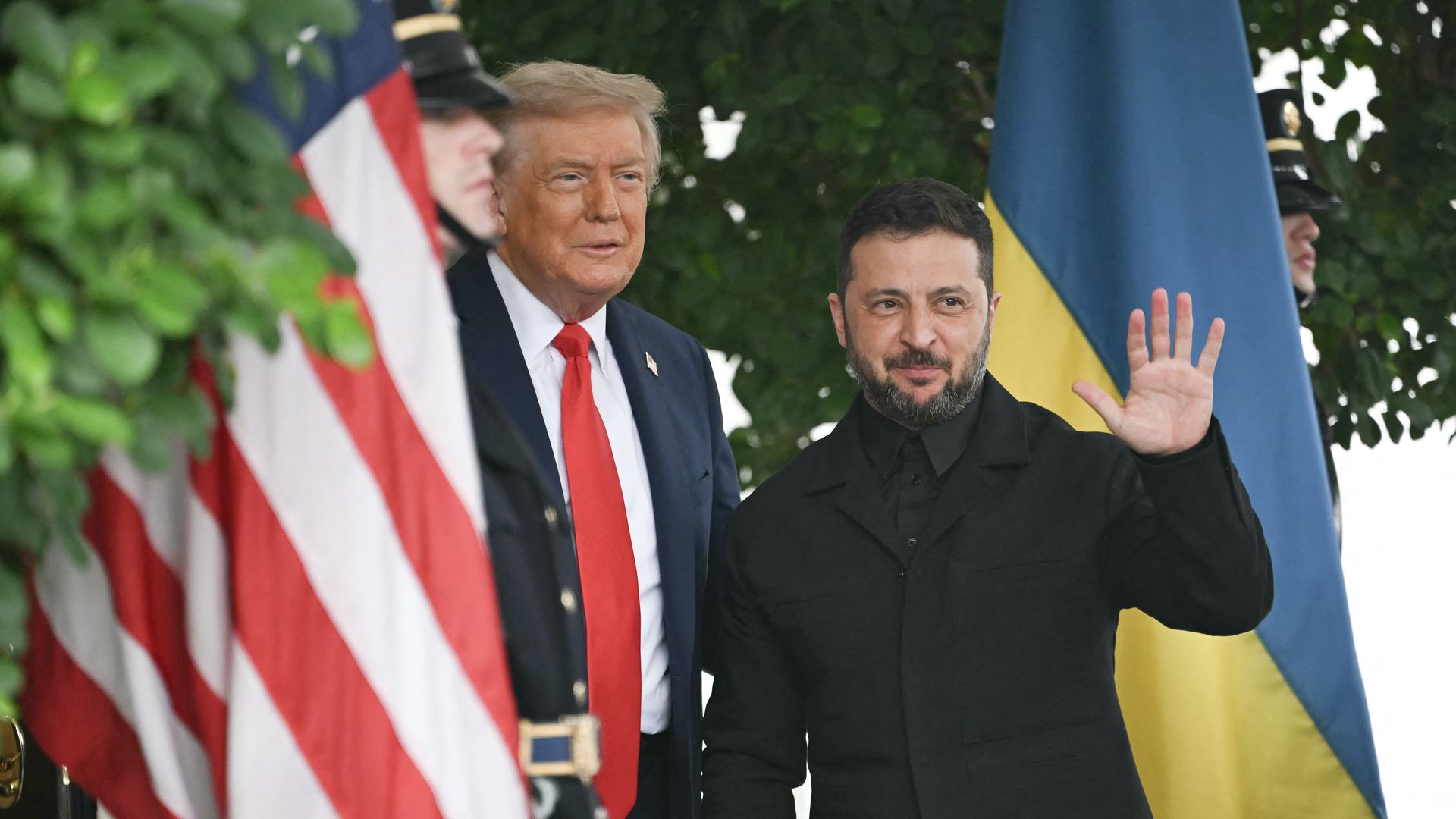 Two men stand side by side outdoors. One man wears a dark suit and red tie, the other a black jacket, waving. U.S. and Ukrainian flags and two uniformed guards are visible in the background.