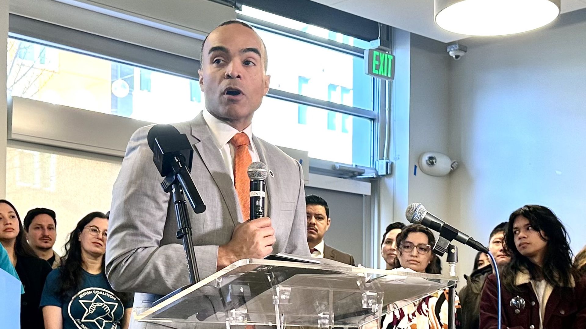 Nick Brown speaks at a podium while holding a microphone while surrounded by people in the background at a press conference.