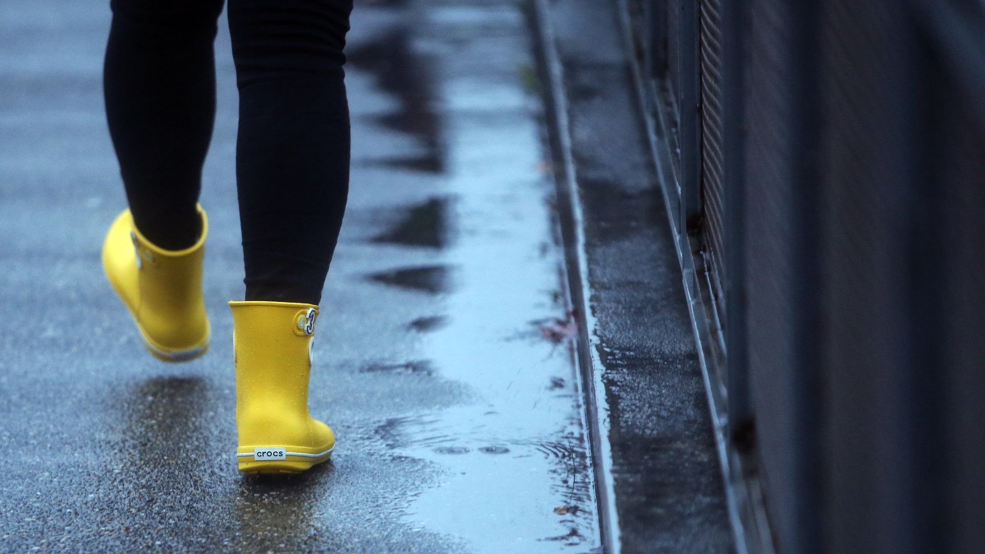 A photo of the legs of a person wearing yellow boots and black leggings on a rain-slicked sidewalk. 