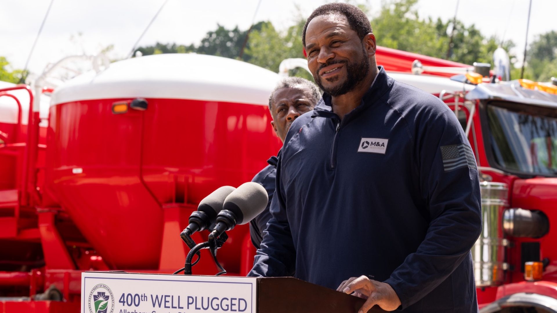 Smiling man in a navy jacket speaks at a podium with microphones at a press event, red fire trucks behind him, and a sign reads "400th WELL PLUGGED, Allegheny County, PA".