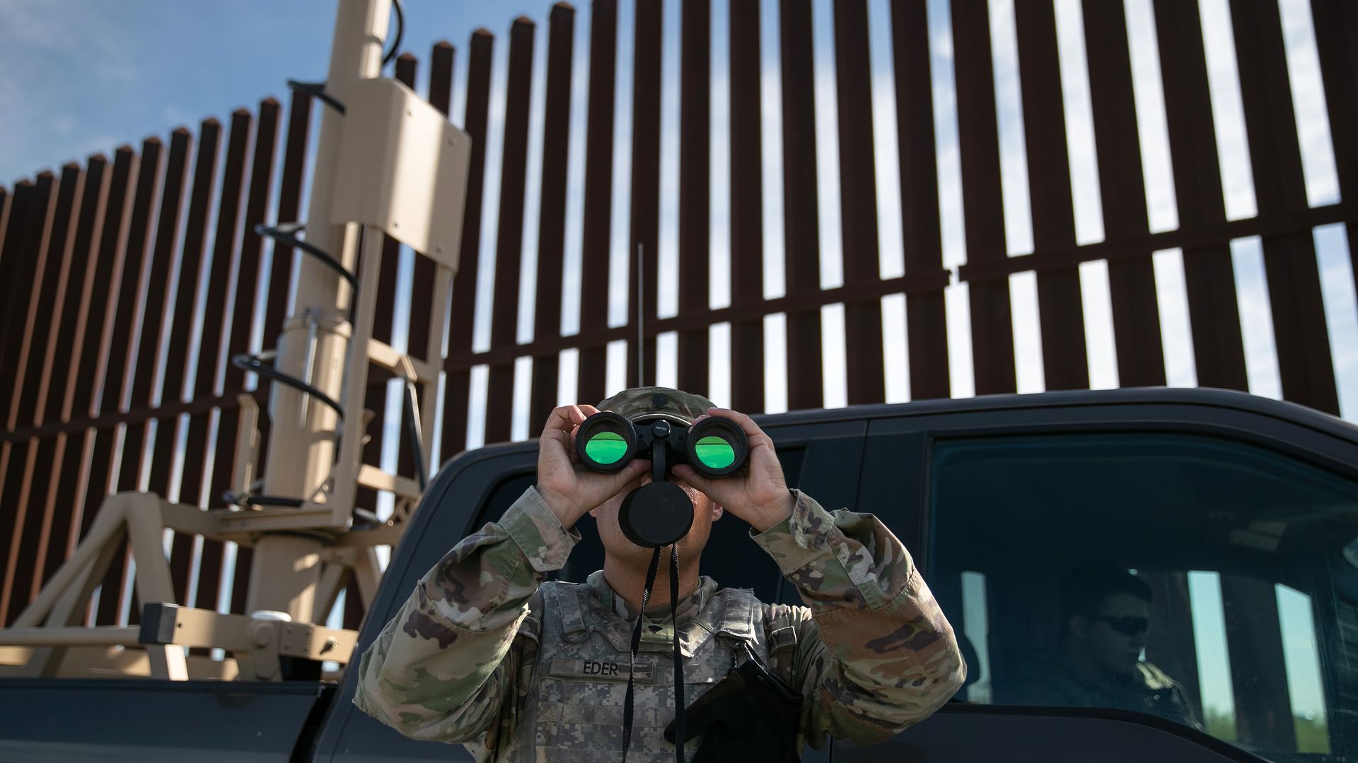In this image, a member of the U.S. army in standard military fatigues holds up binoculars to his face while standing in front of a pickup truck at a tall border wall fence.