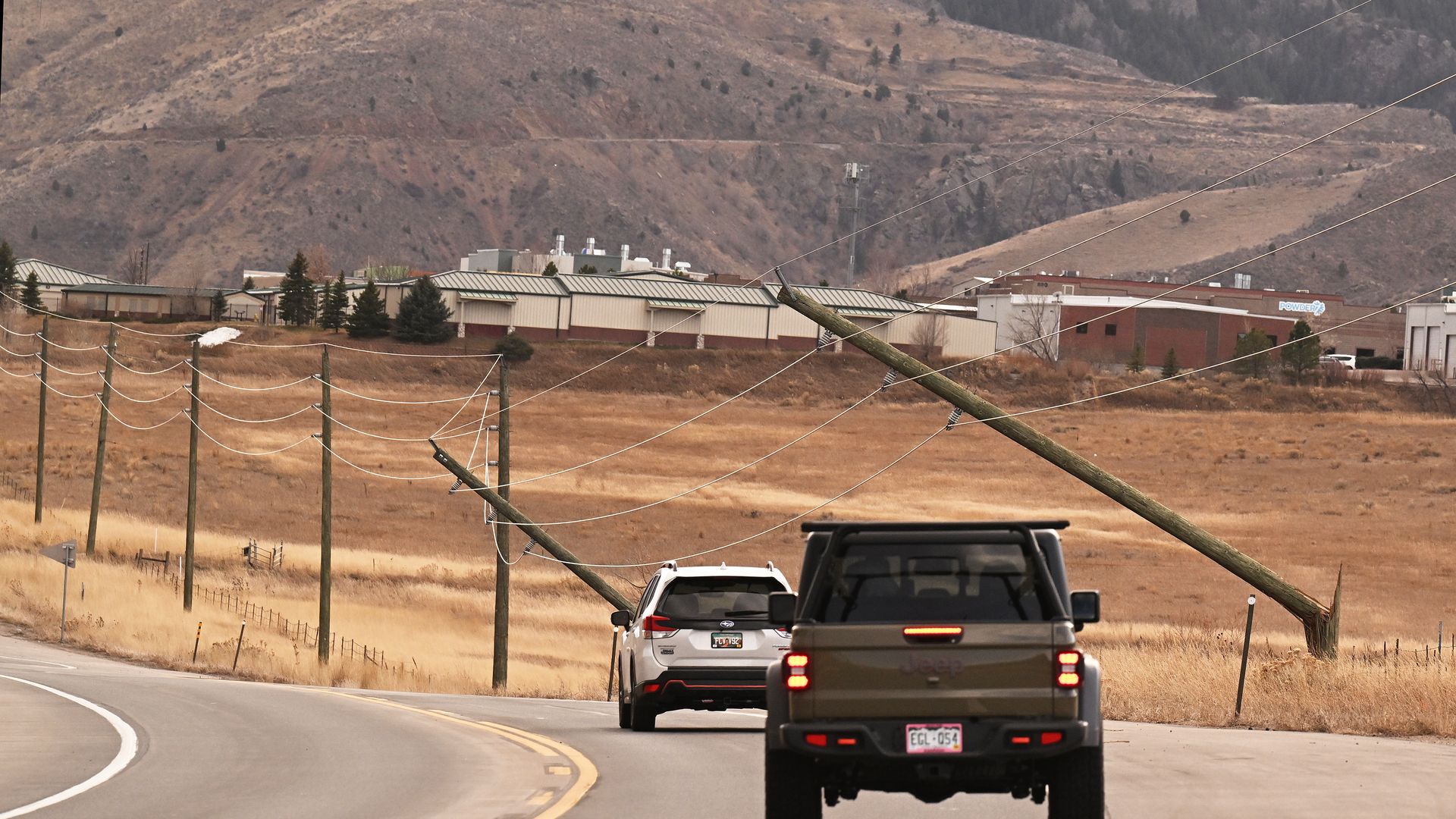 Two vehicles drive past leaning utility poles and sagging power lines along a curved rural road with dry grass and brown hills in the background under an overcast sky.