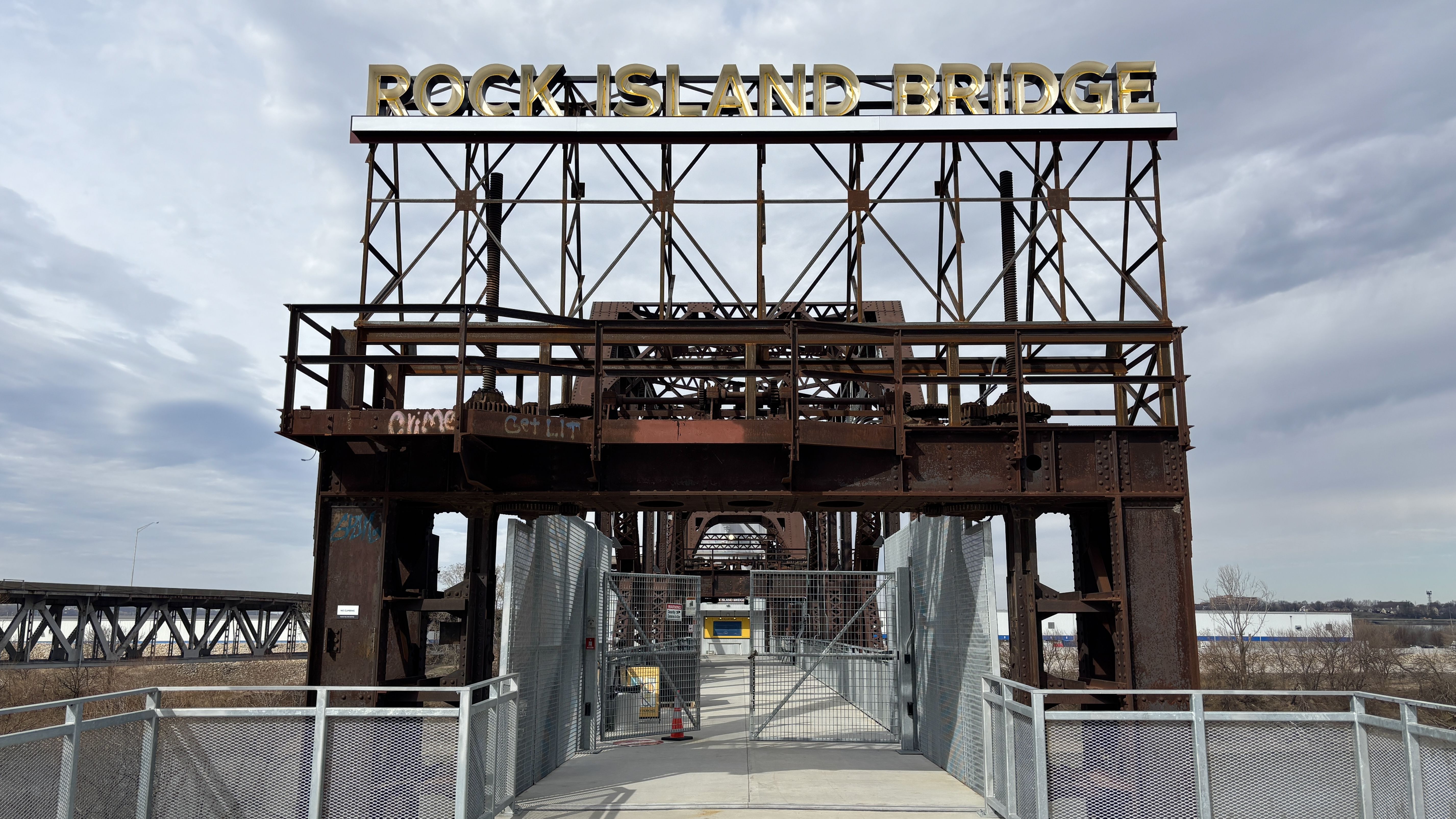 Rusty iron bridge frame with a gold sign reading "ROCK ISLAND BRIDGE" atop a lattice structure. A fenced walkway passes through gates toward a distant, cloudy sky.