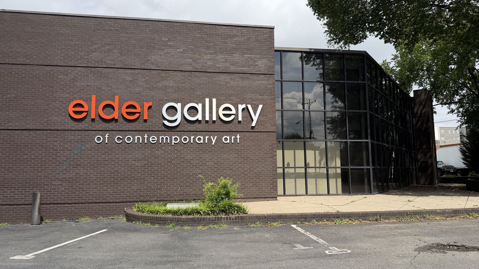 Exterior brick gallery building with large glass windows. Prominent orange elder sign and white gallery text, with smaller of contemporary art underneath; trees and a parking lot nearby.