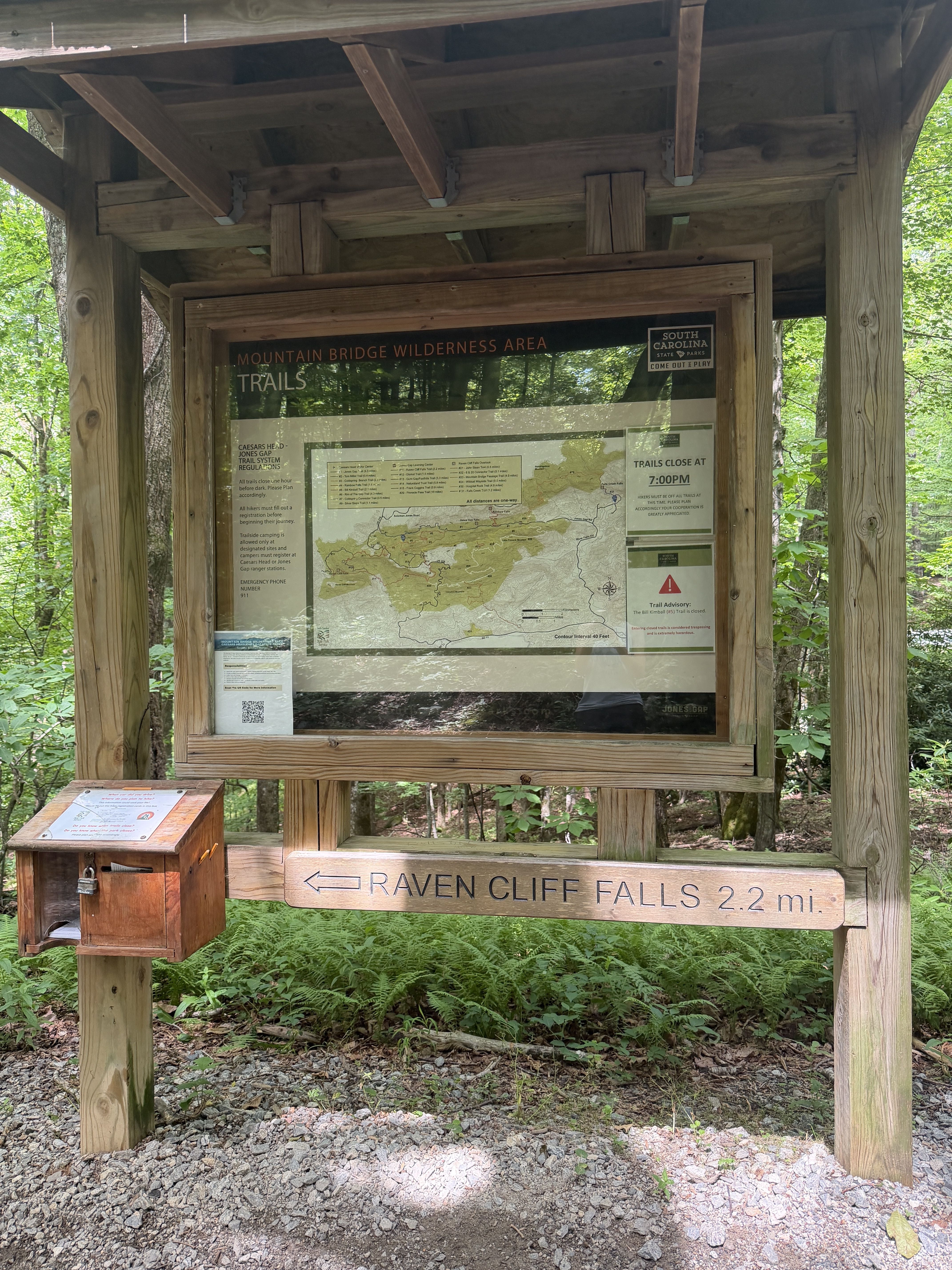Rustic wooden trail information kiosk in a lush forest. A glass-front map shows Mountain Bridge Wilderness Area trails; bottom beam reads Raven Cliff Falls 2.2 mi, with a lockbox nearby.