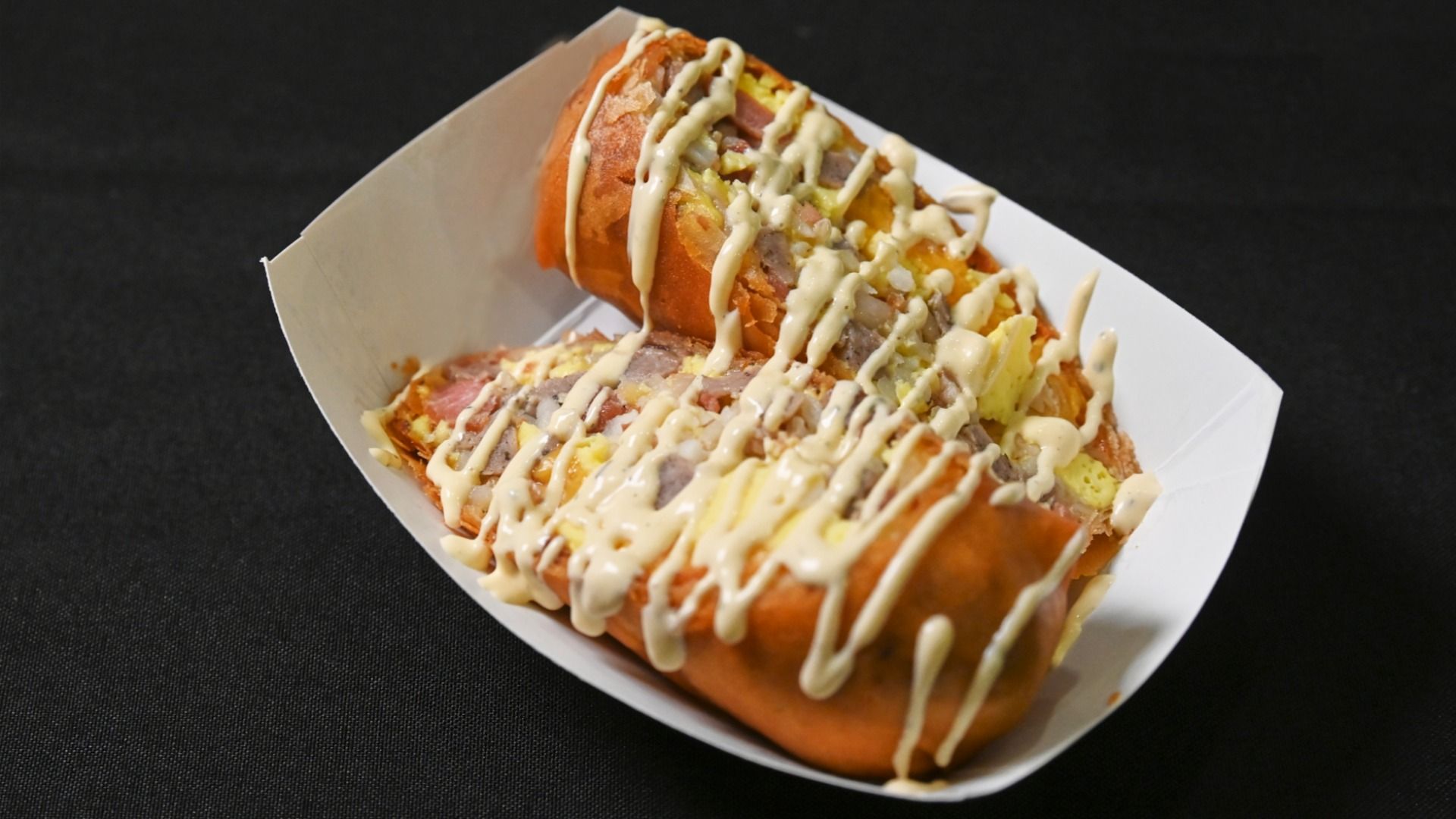 Two loaded hot dogs in a white tray, topped with corn, onions, and meat, with a zigzag of pale sauce on a dark background.