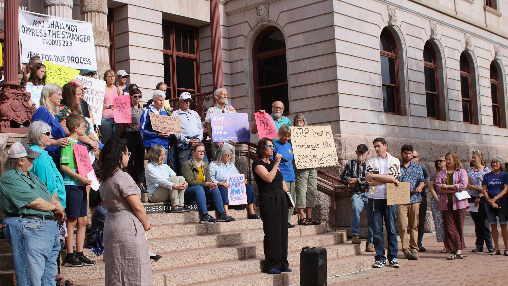 Diverse group of people on the stone steps of a grand building with arched windows, holding protest signs about immigration and human rights; a large white banner is visible.