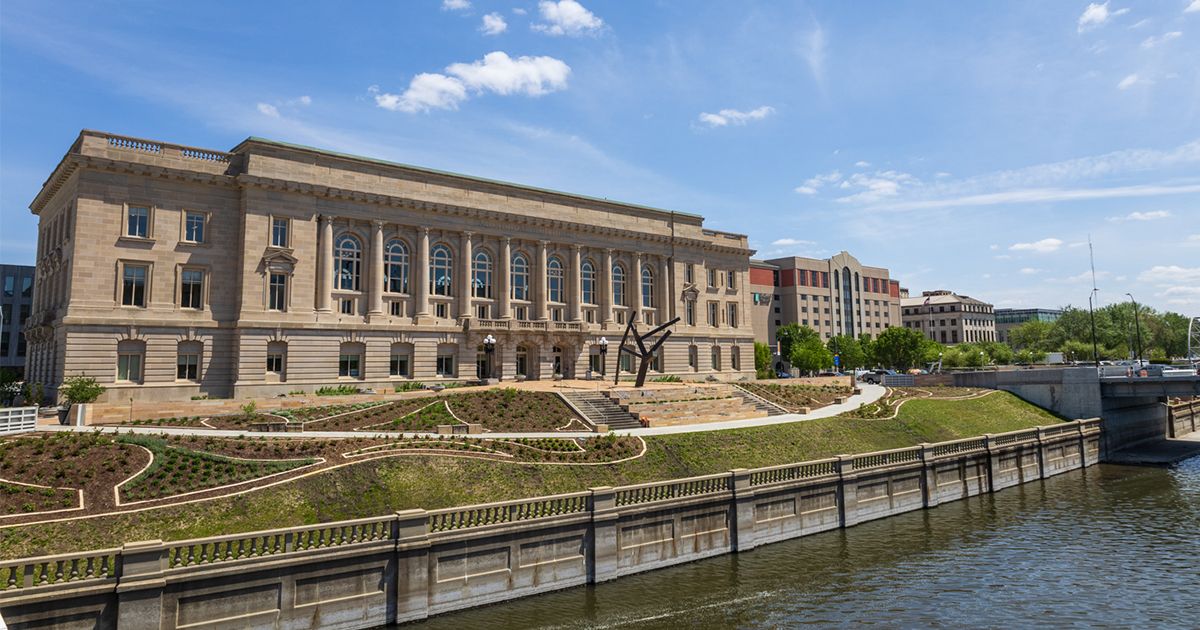 Large beige classical building with tall arched windows beside a river; blue sky with clouds above; landscaped area with paths and a sculpture in front; nearby modern buildings and a bridge.