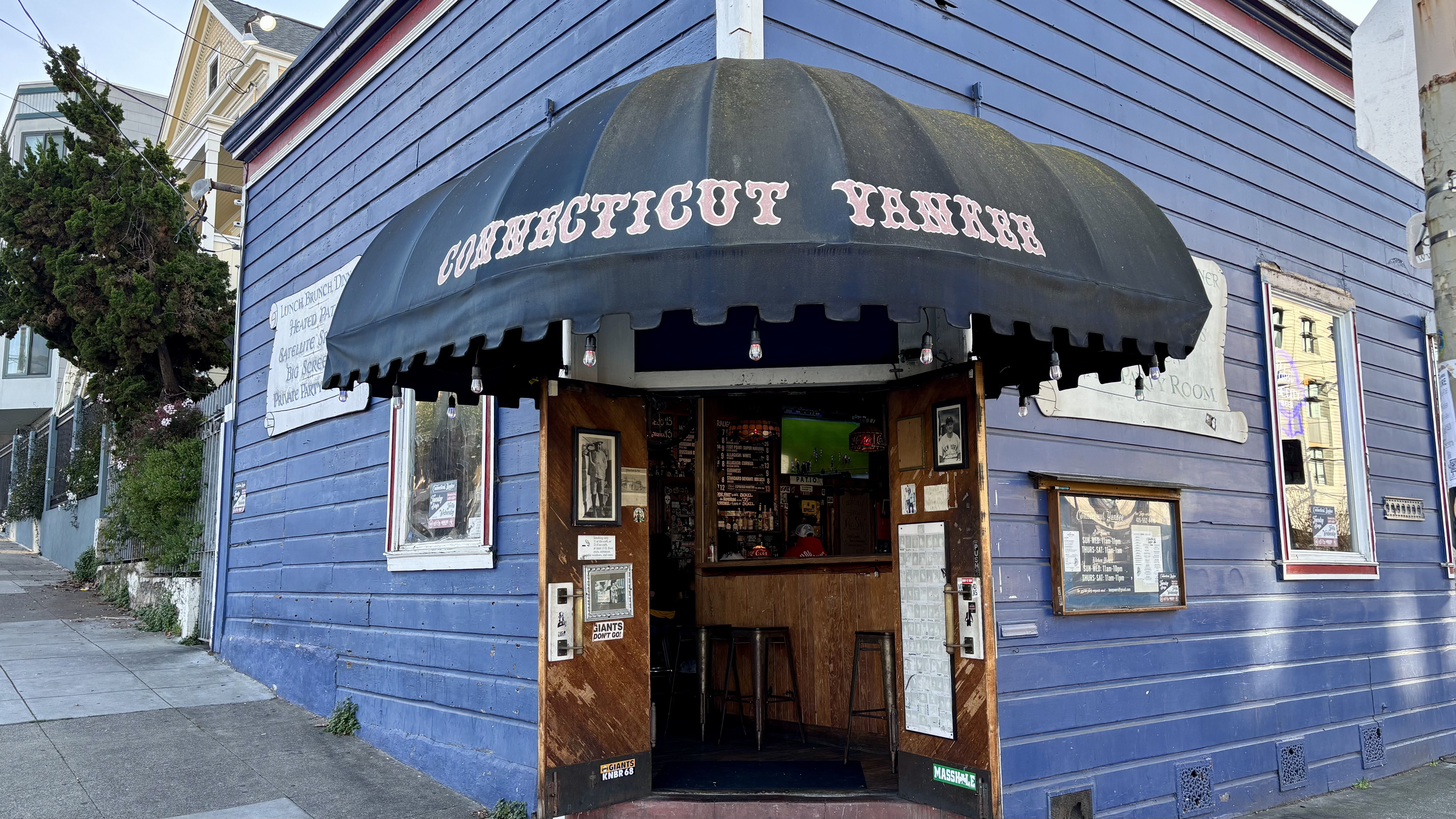 Blue wooden building with black awning reading "CONNECTICUT YANKEE," open entrance showing bar stools inside, posters, and a TV with a sports game on.