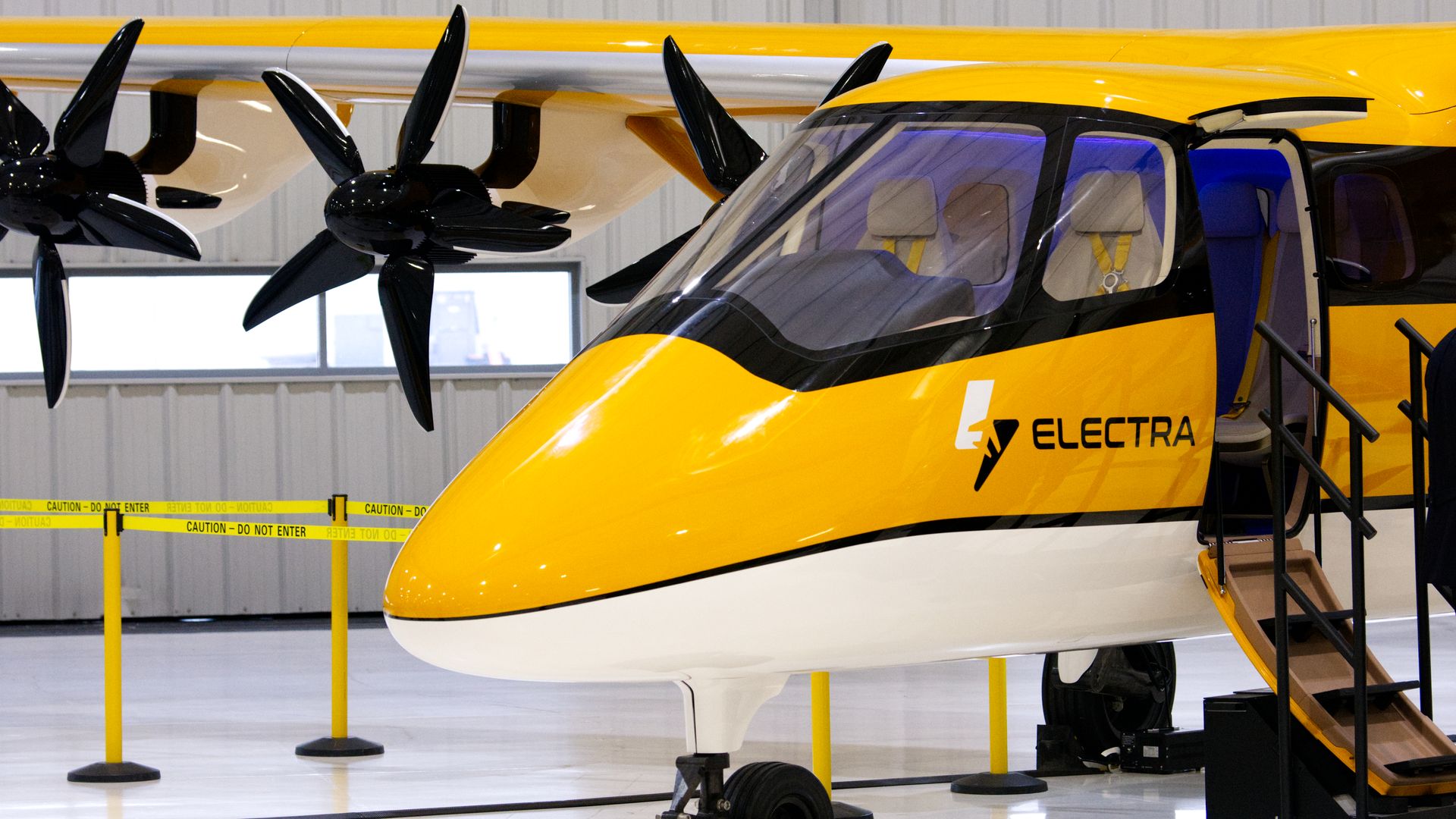Yellow and white Electra aircraft in a hangar with open side door, black propellers, yellow caution tape surrounding it, and stairs leading into the cabin.