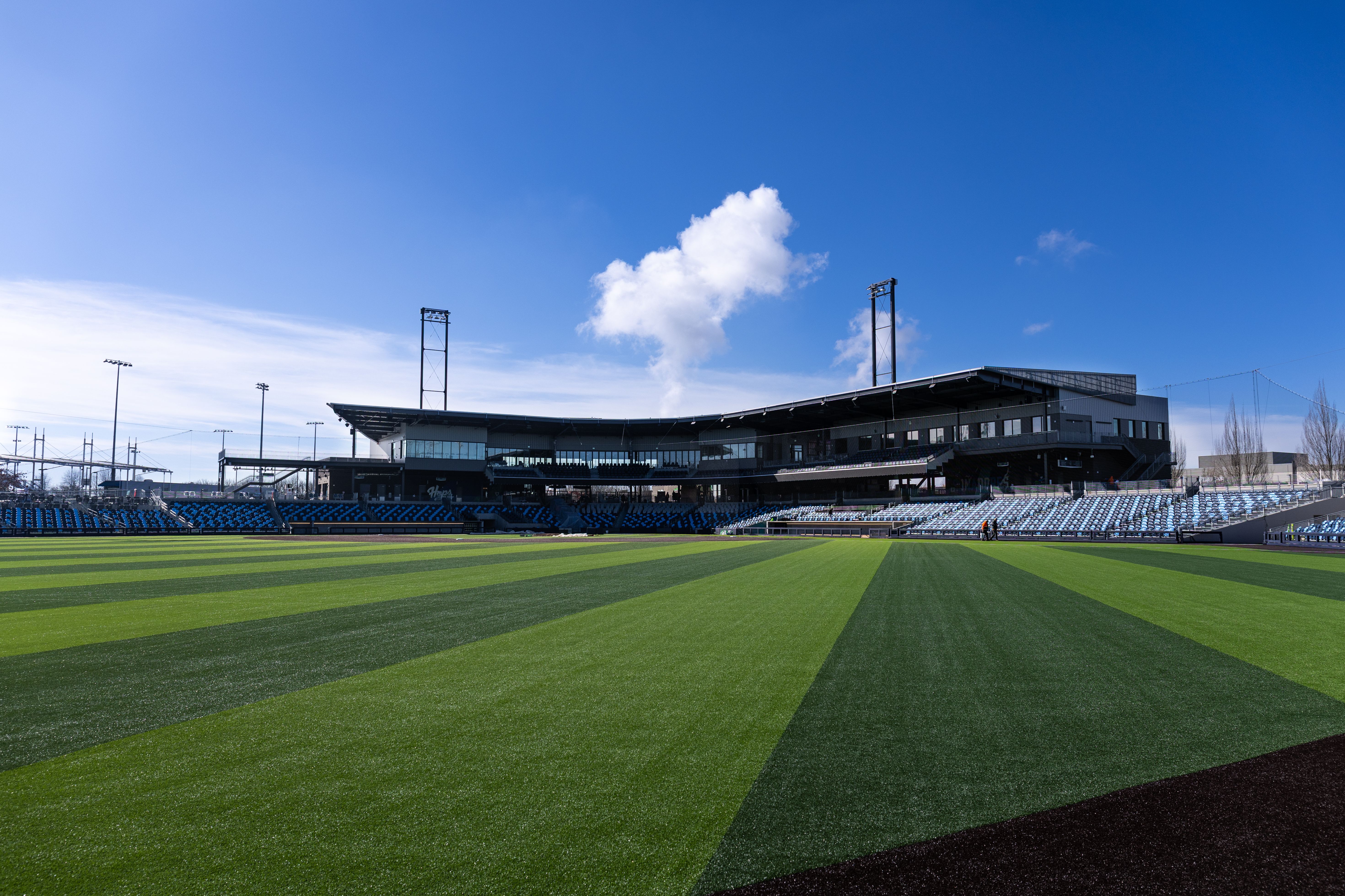 Wide view of a modern stadium with a dark, multi-level grandstand and blue seating, and a striped green field under a clear blue sky with a few white clouds.