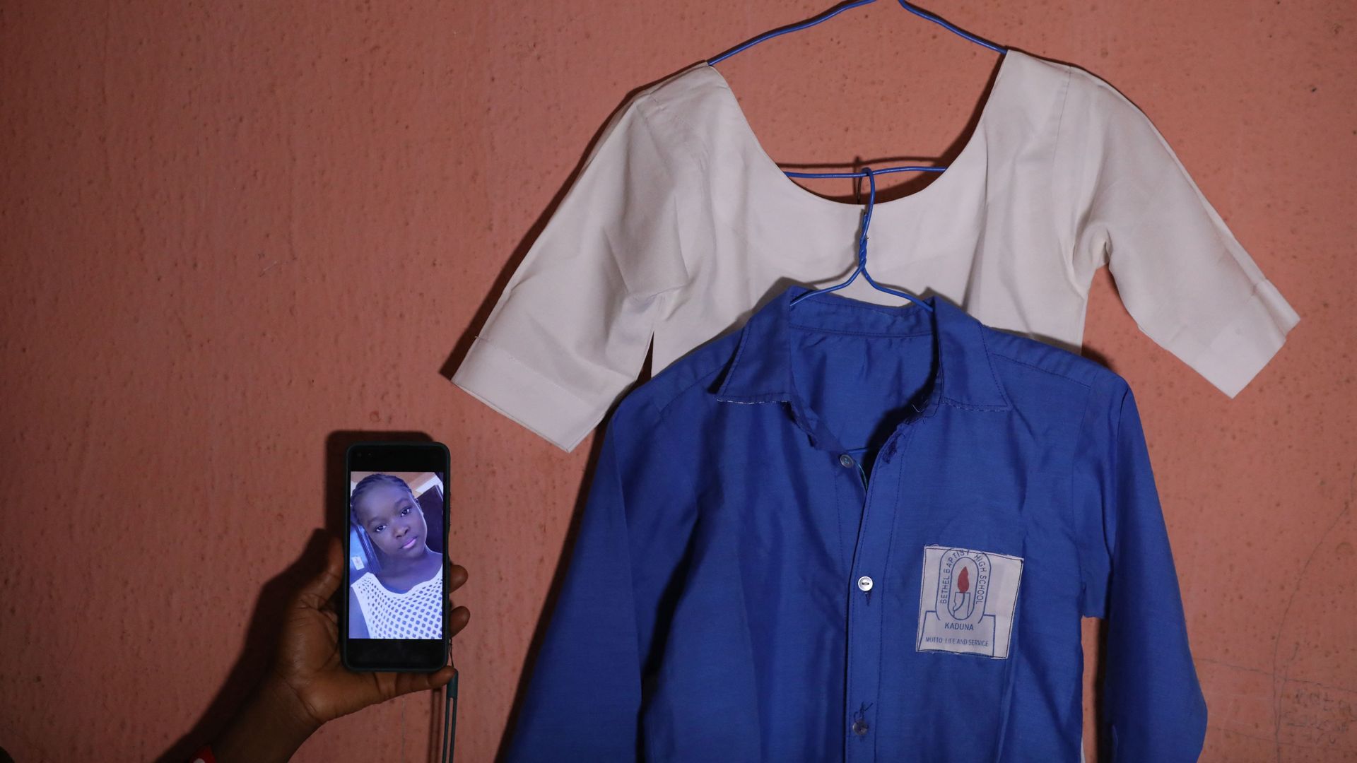 A hand holds a phone with a picture of a girl next to a hanging school uniform